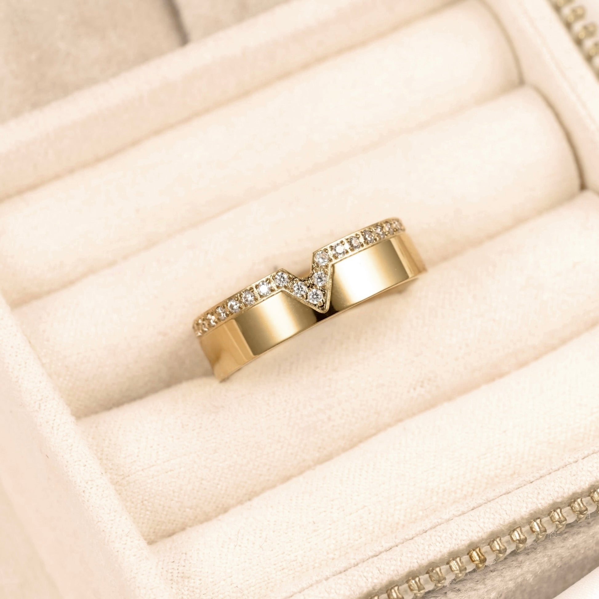 A gold wedding band with a chevron design paved with small clear stones, displayed on a cream-colored velvet ring box.