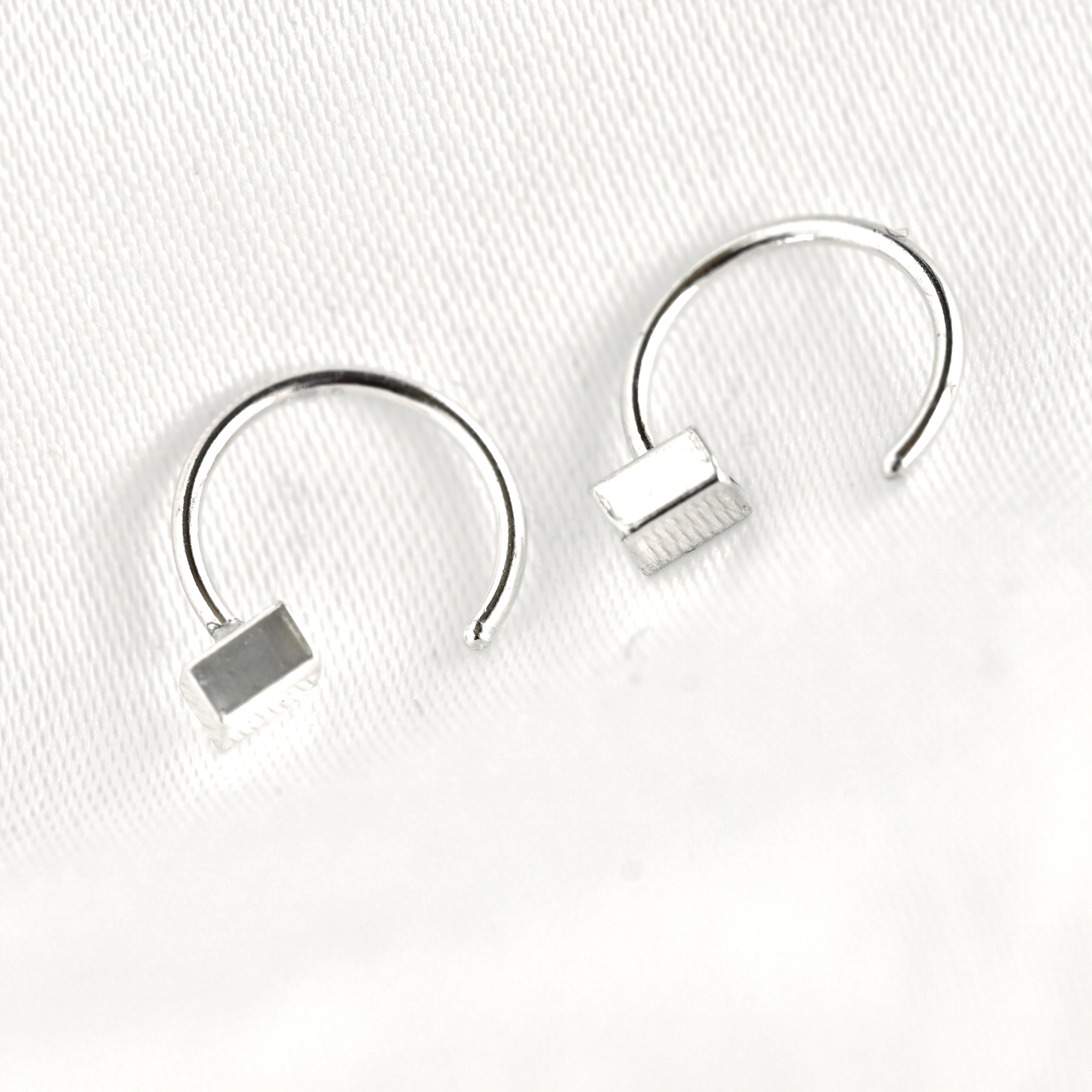 A pair of silver-toned open hoop earrings with a small rectangular block detail at the end, displayed on a textured white surface.