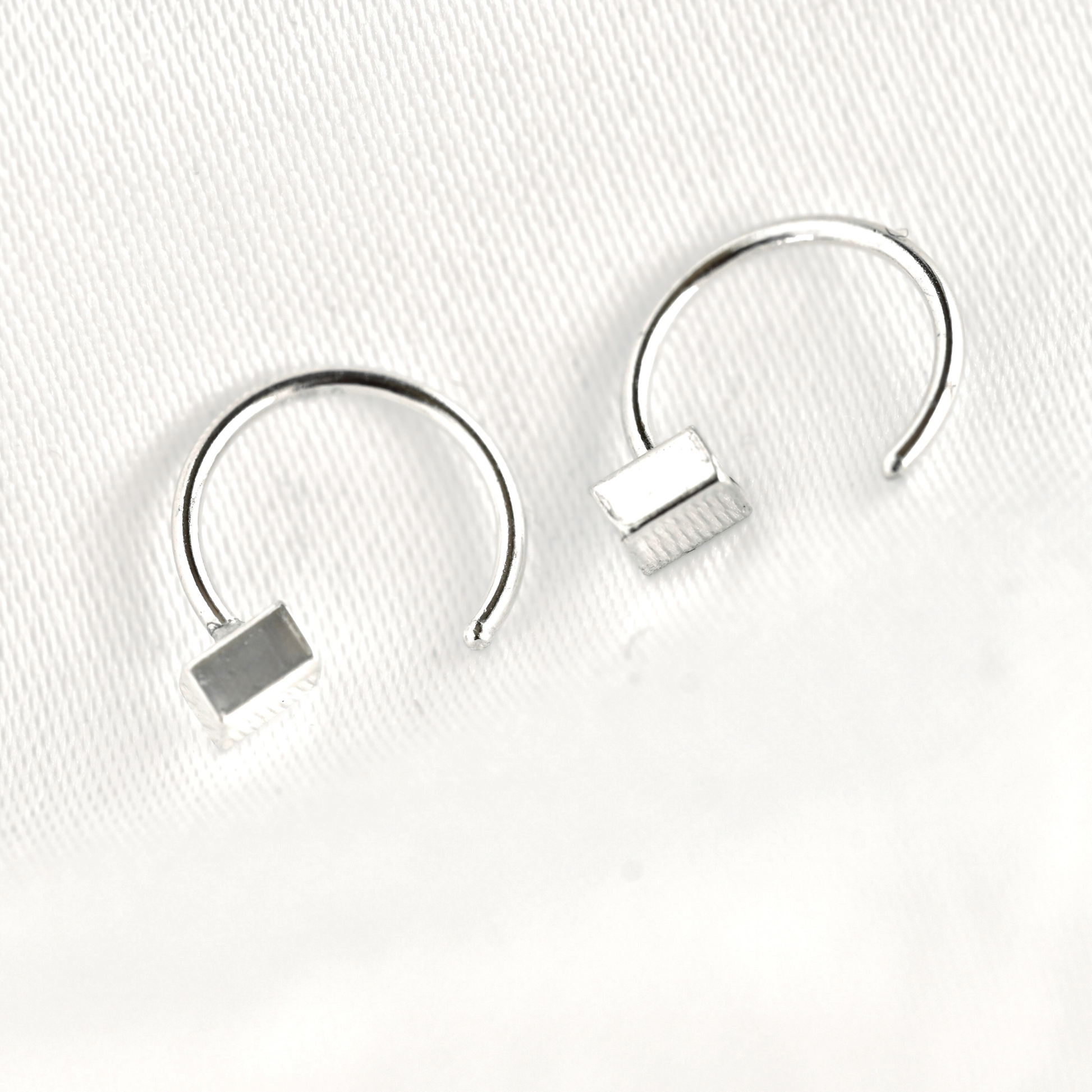 A pair of silver-toned open hoop earrings with a small rectangular block detail at the end, displayed on a textured white surface.