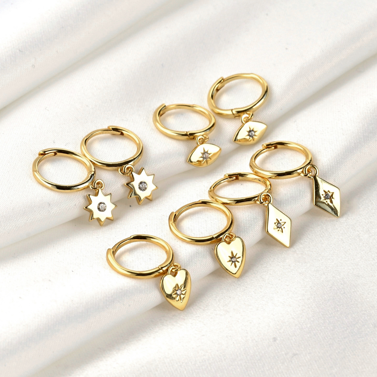 Set of gold earrings with various charms inclduing star, evil eye, heart and rhombus with a tiny CZ in the center on a white background
