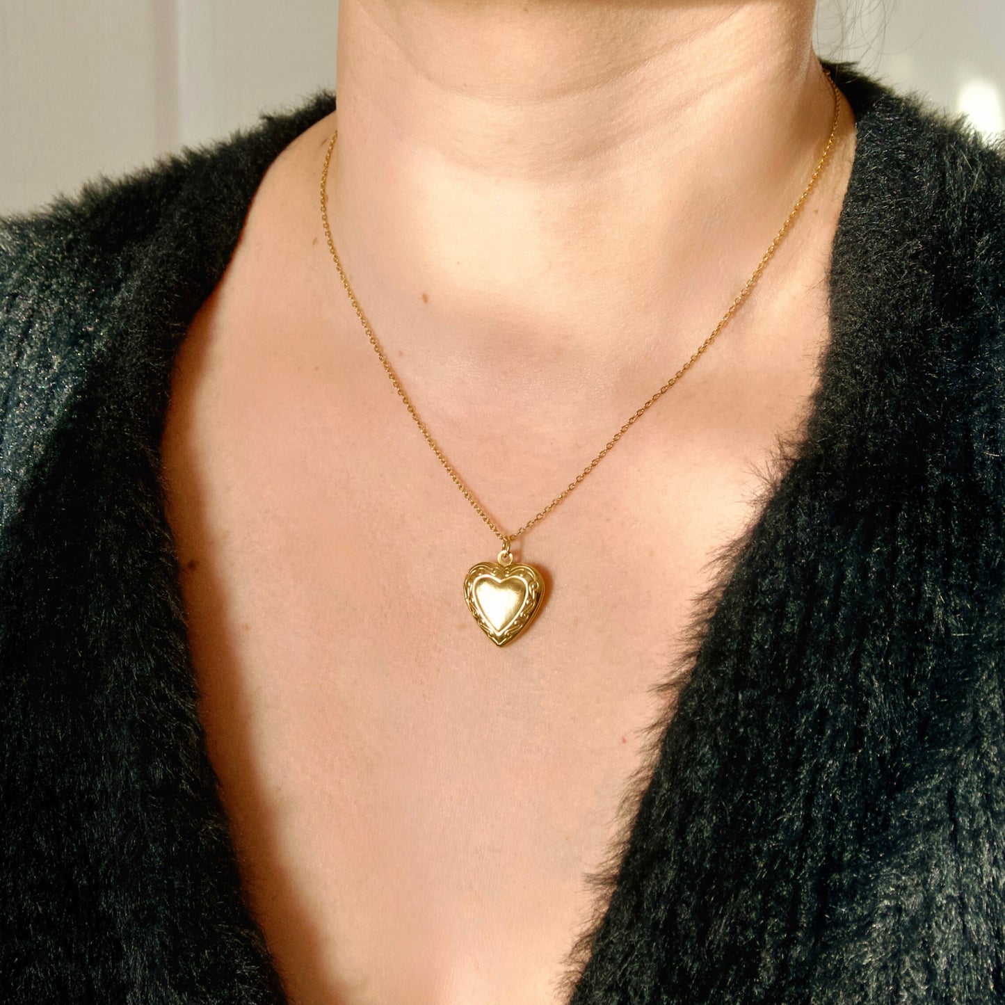 Gold heart locket necklace worn by a person with a black textured garment.
