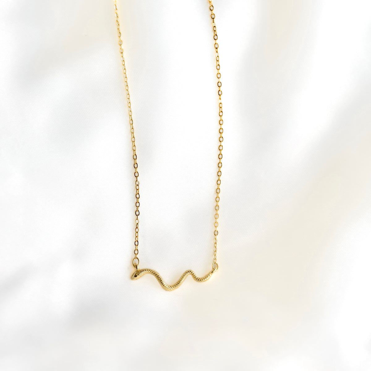 A delicate gold chain necklace with a small, wavy snake pendant, displayed against a soft, white marble background.