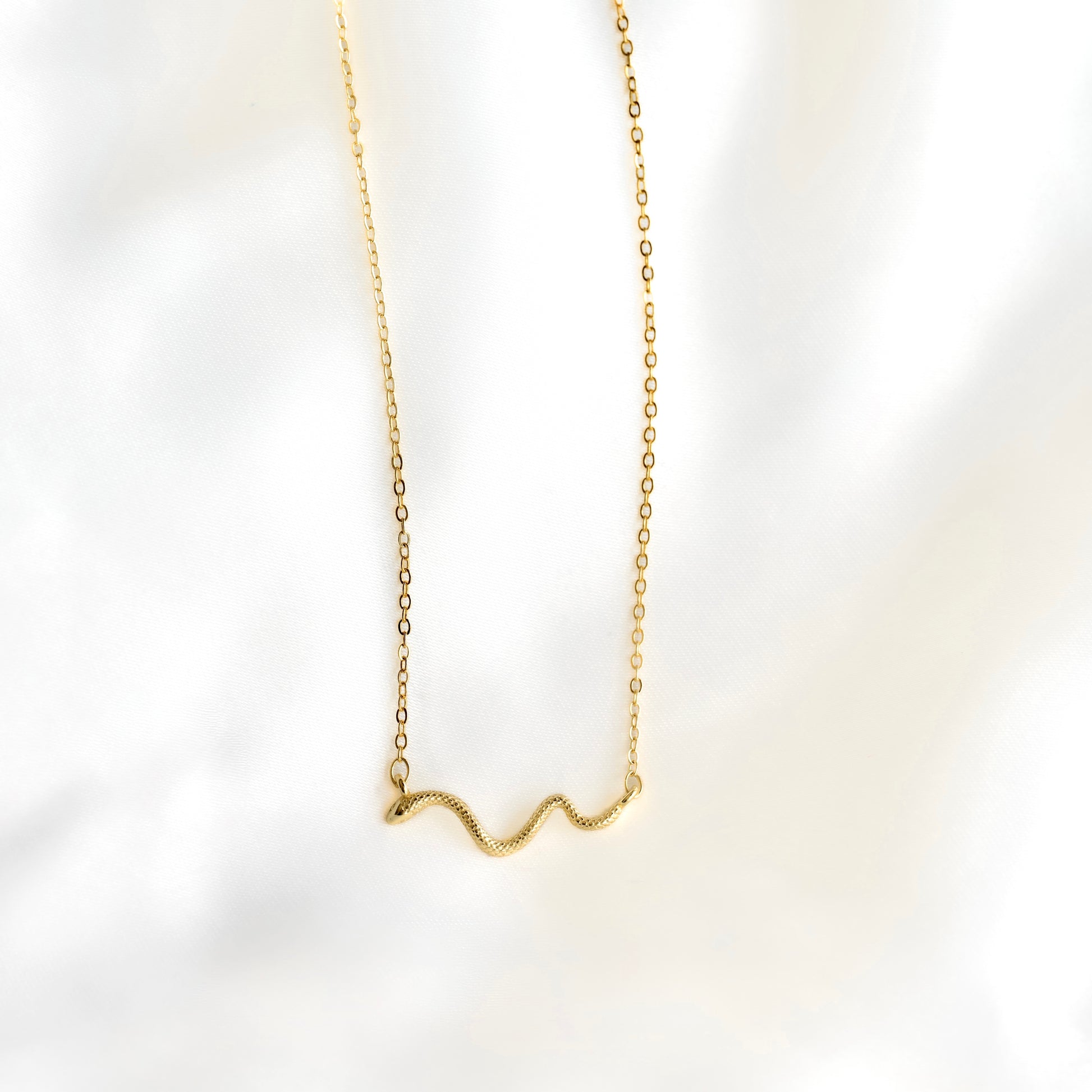 A delicate gold chain necklace with a small, wavy snake pendant, displayed against a soft, white marble background.