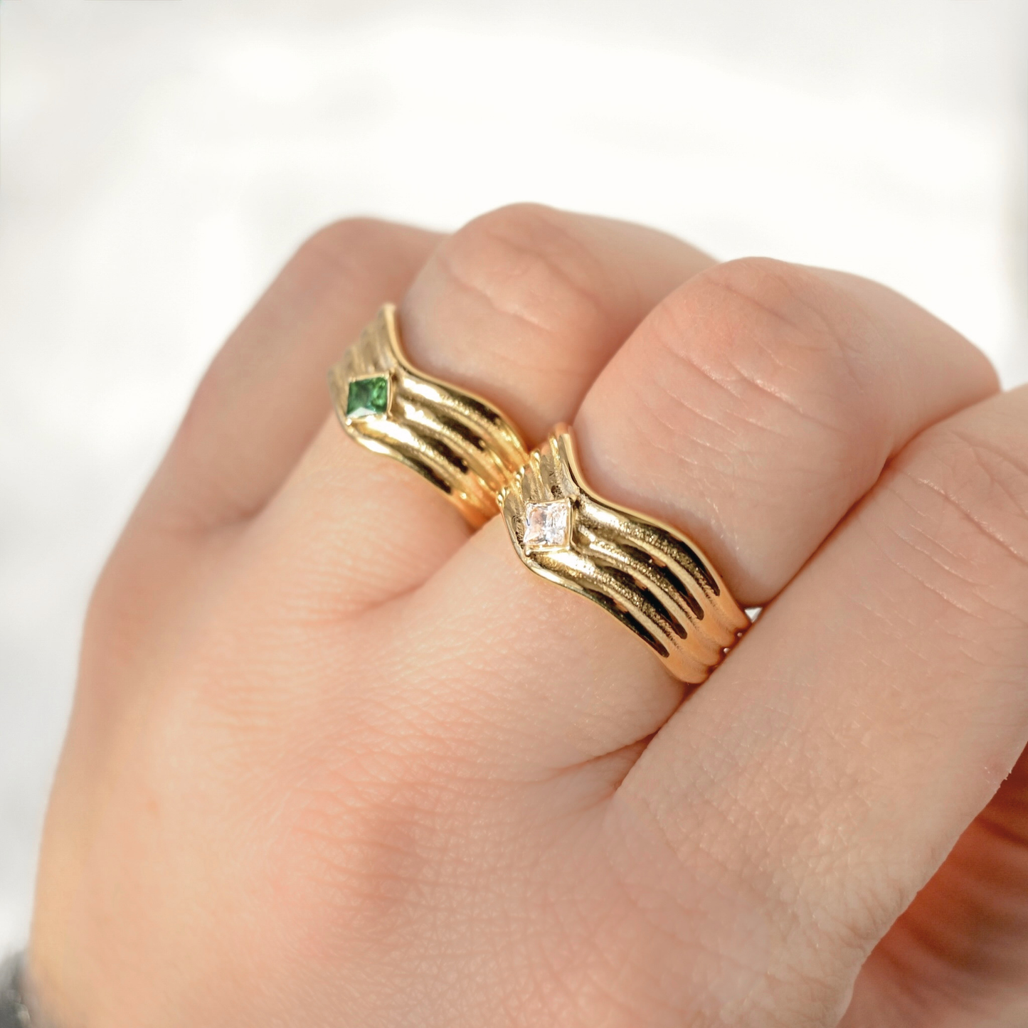 A close-up of a hand wearing two gold rings with a wing-like design, one featuring a small green stone and the other a small clear stone.