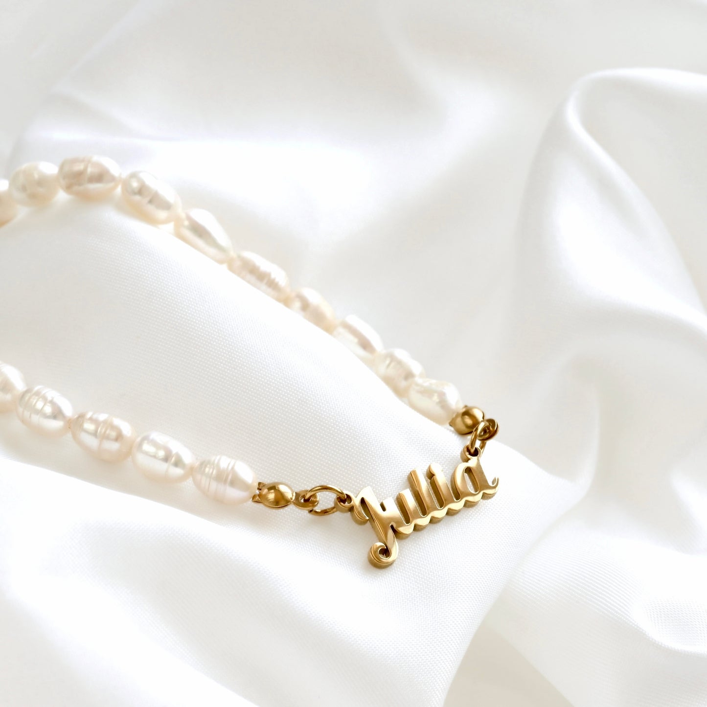 A personalized pearl necklace with a gold nameplate charm spelling 'Julia', displayed on a draped white satin fabric.