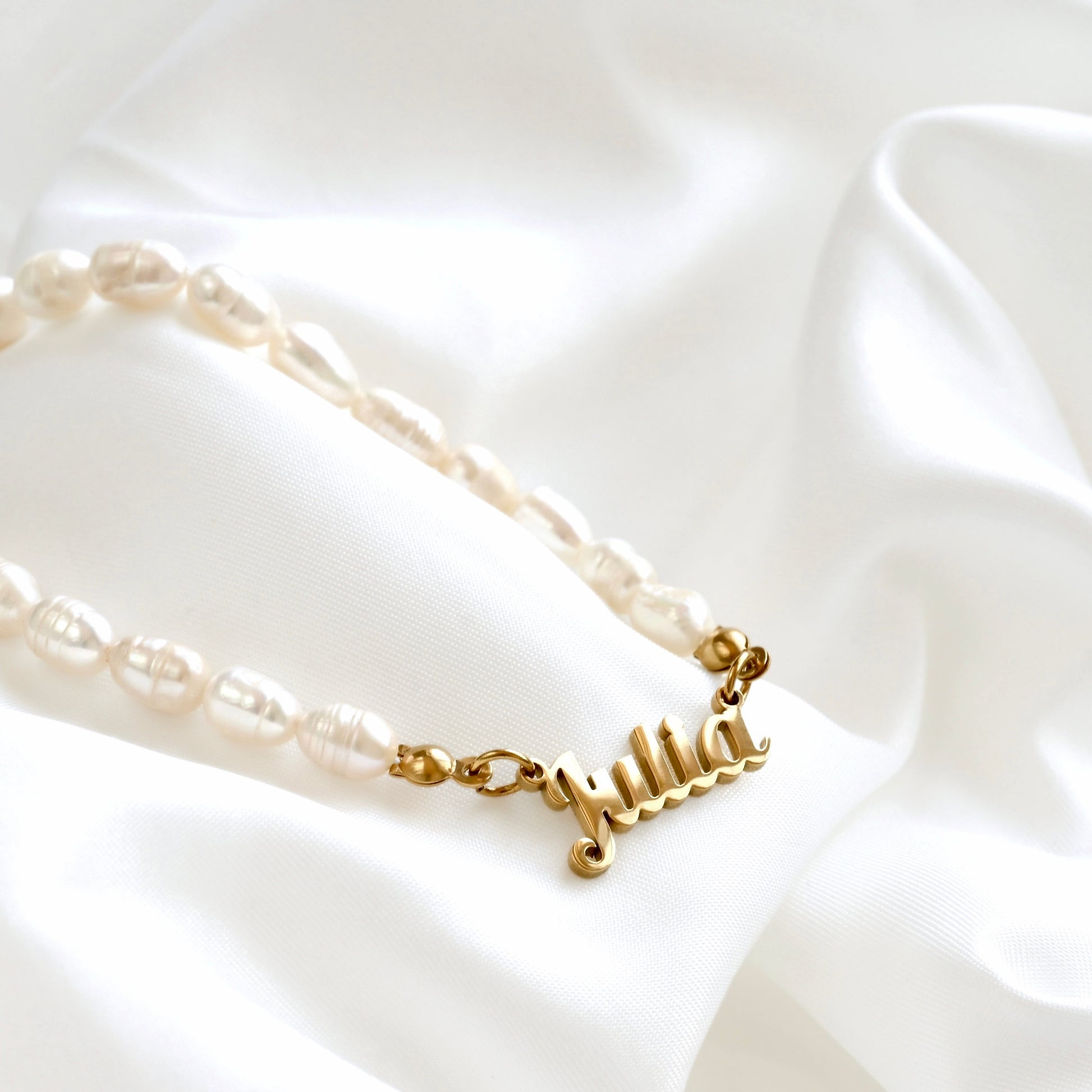 A personalized pearl necklace with a gold nameplate charm spelling 'Julia', displayed on a draped white satin fabric.
