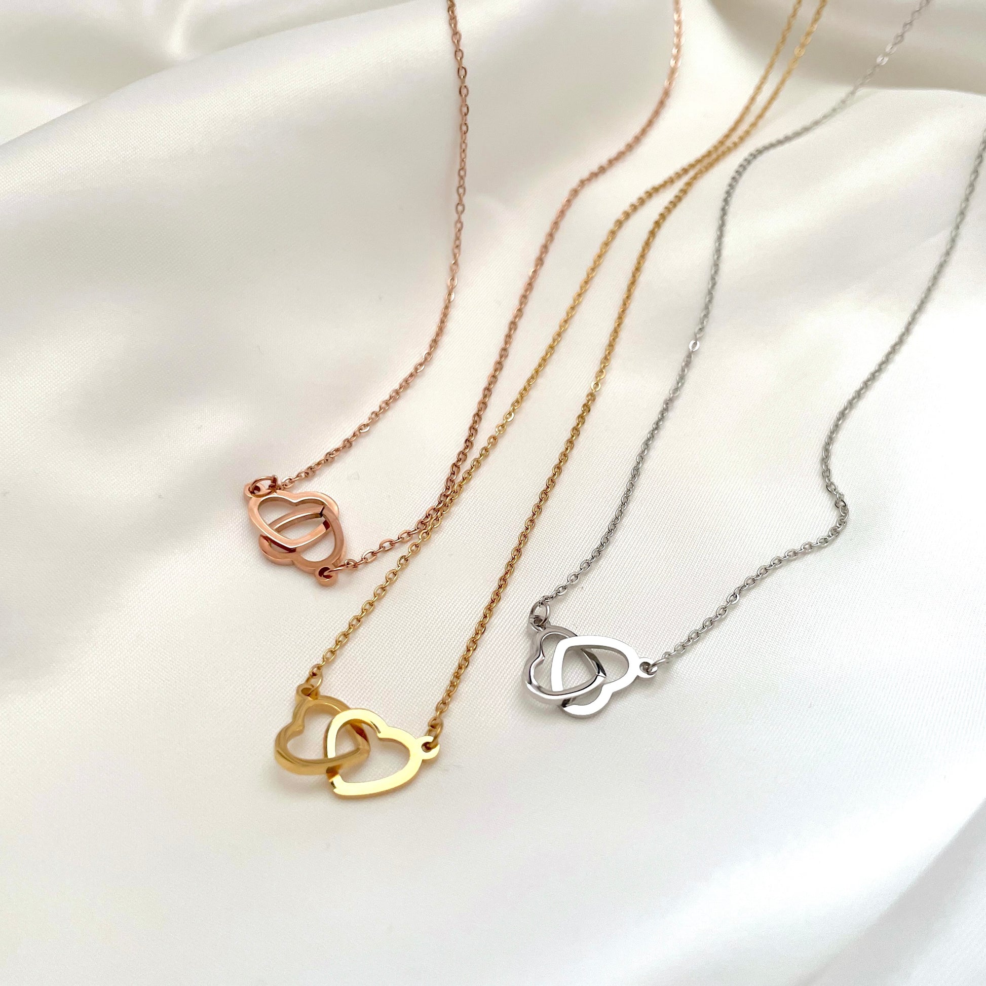 Three necklaces with interlocking heart pendants in gold, rose gold and silver on a white surface