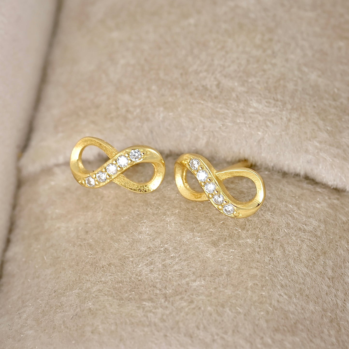 A pair of gold infinity-shaped stud earrings set with small clear stones, displayed on a beige textured velvet jewelry surface.