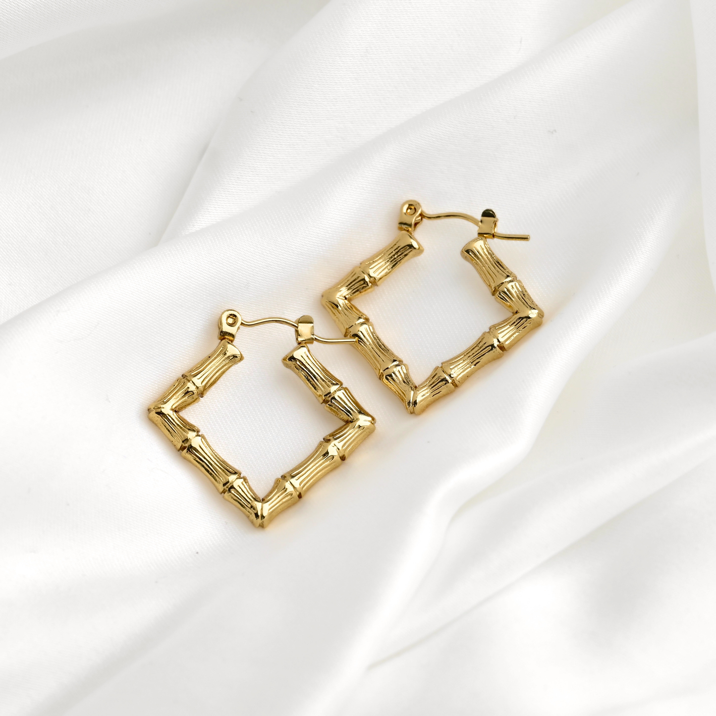 A pair of gold  square hoop earrings designed to look like bamboo stalks, resting on a draped white satin fabric background.