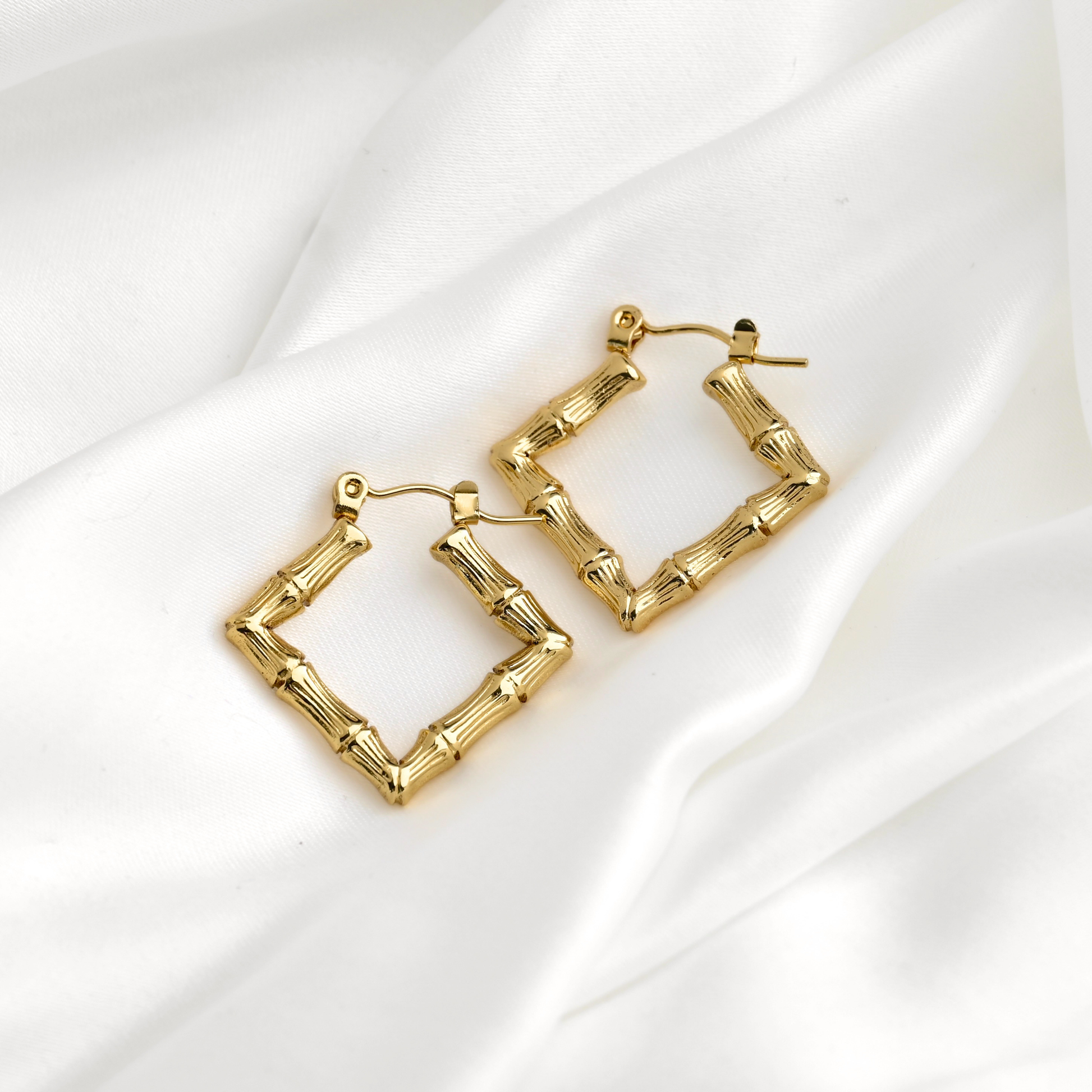 A pair of gold  square hoop earrings designed to look like bamboo stalks, resting on a draped white satin fabric background.