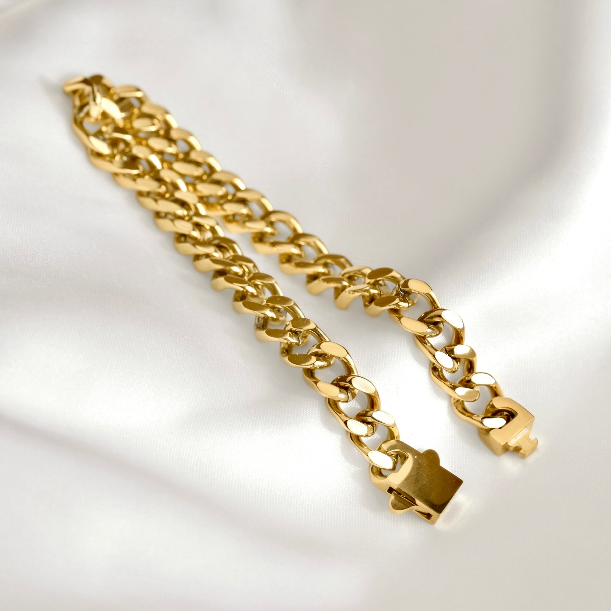 A gold Cuban link chain bracelet with a polished finish, displayed on a draped white satin fabric background.