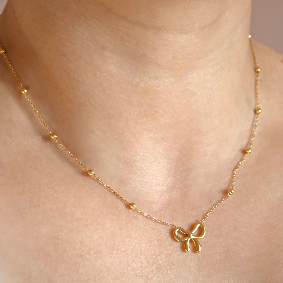 A delicate gold chain necklace with small bead accents and a bow-shaped pendant, shown worn on a person's neck.