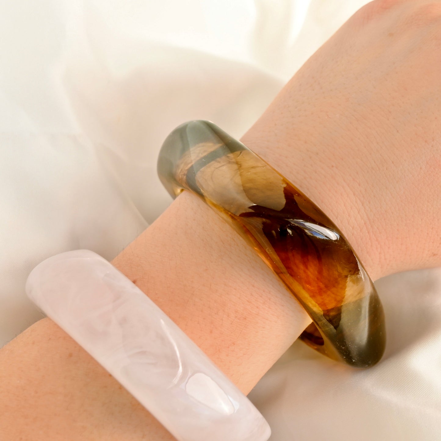 A close-up of a hand wearing a thick, polished translucent brown bangle bracelet, with a second pale translucent bangle partially visible underneath on a soft white fabric background.