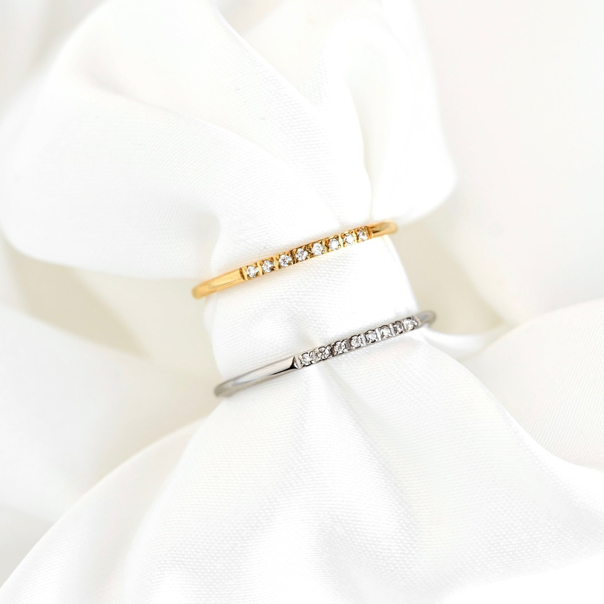 Two slim rings displayed on white fabric: one gold and one silver band with a row of small clear stones.