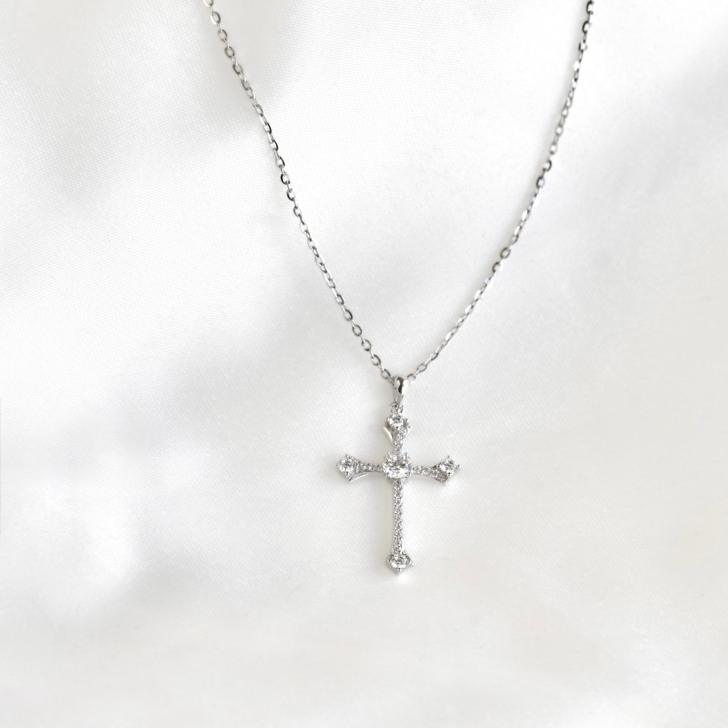 A silver cross pendant necklace with clear stones, displayed on a soft white fabric background.