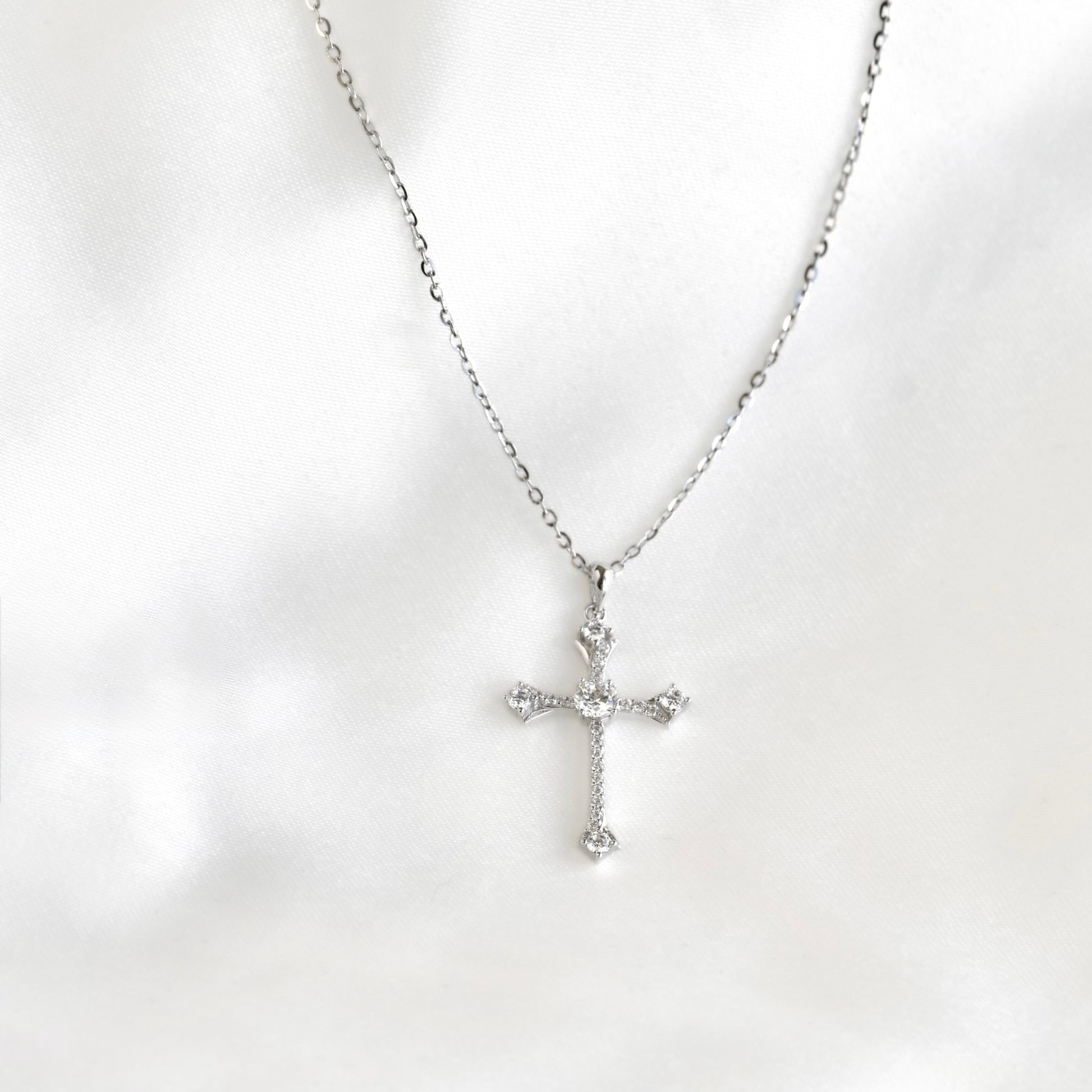 A silver cross pendant necklace with clear stones, displayed on a soft white fabric background.