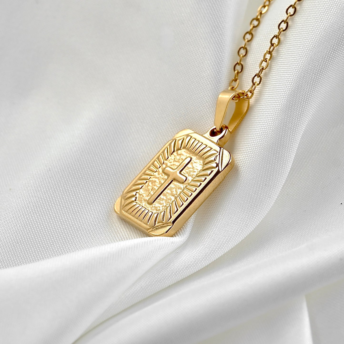 A gold-toned cross tag pendant necklace with an engraved cross and radiating lines, resting on draped white fabric.