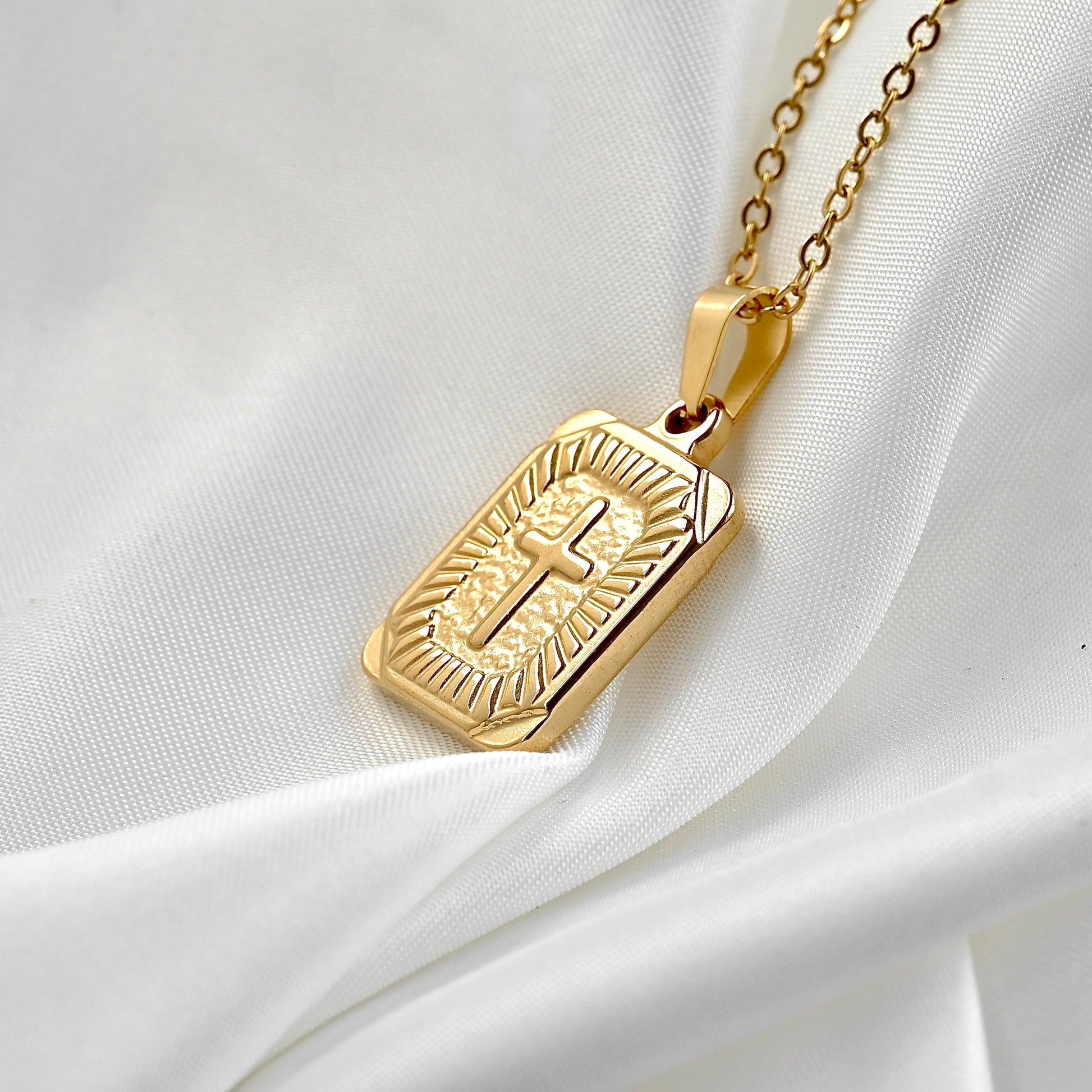 A gold-toned cross tag pendant necklace with an engraved cross and radiating lines, resting on draped white fabric.
