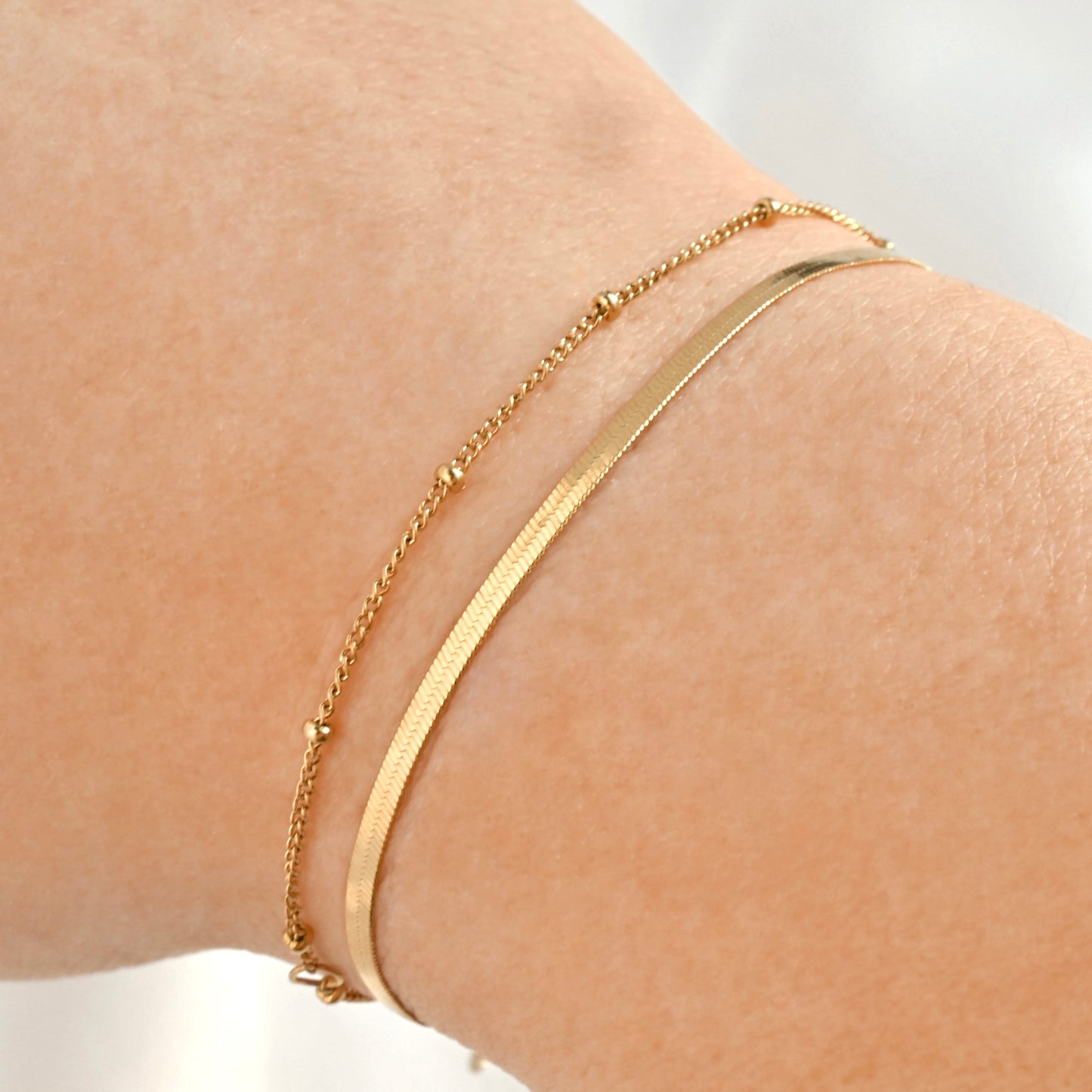 Dainty Gold herringbone bracelet stacked with a satellite chain on a person's wrist against a neutral background
