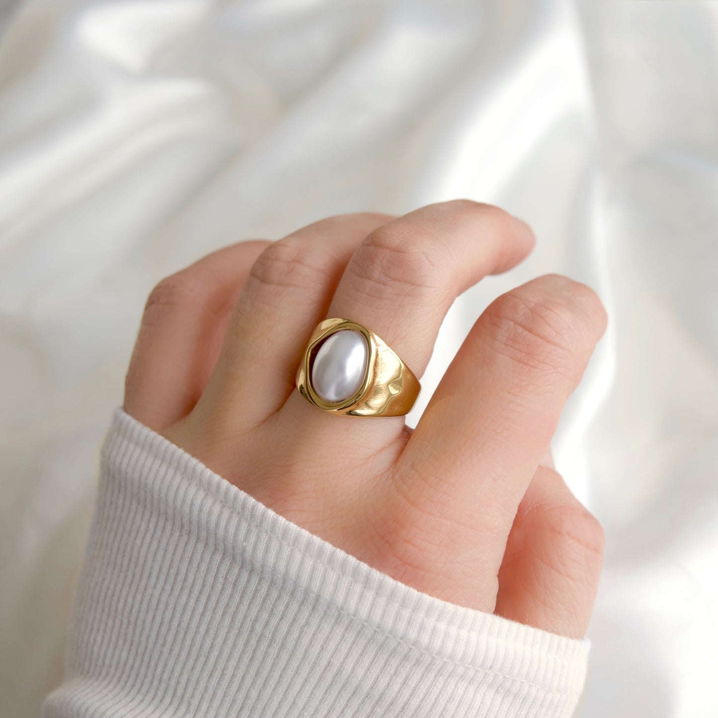 A gold signet-style ring with a large white pearl-like oval center, worn on a person's finger against a soft white fabric background.