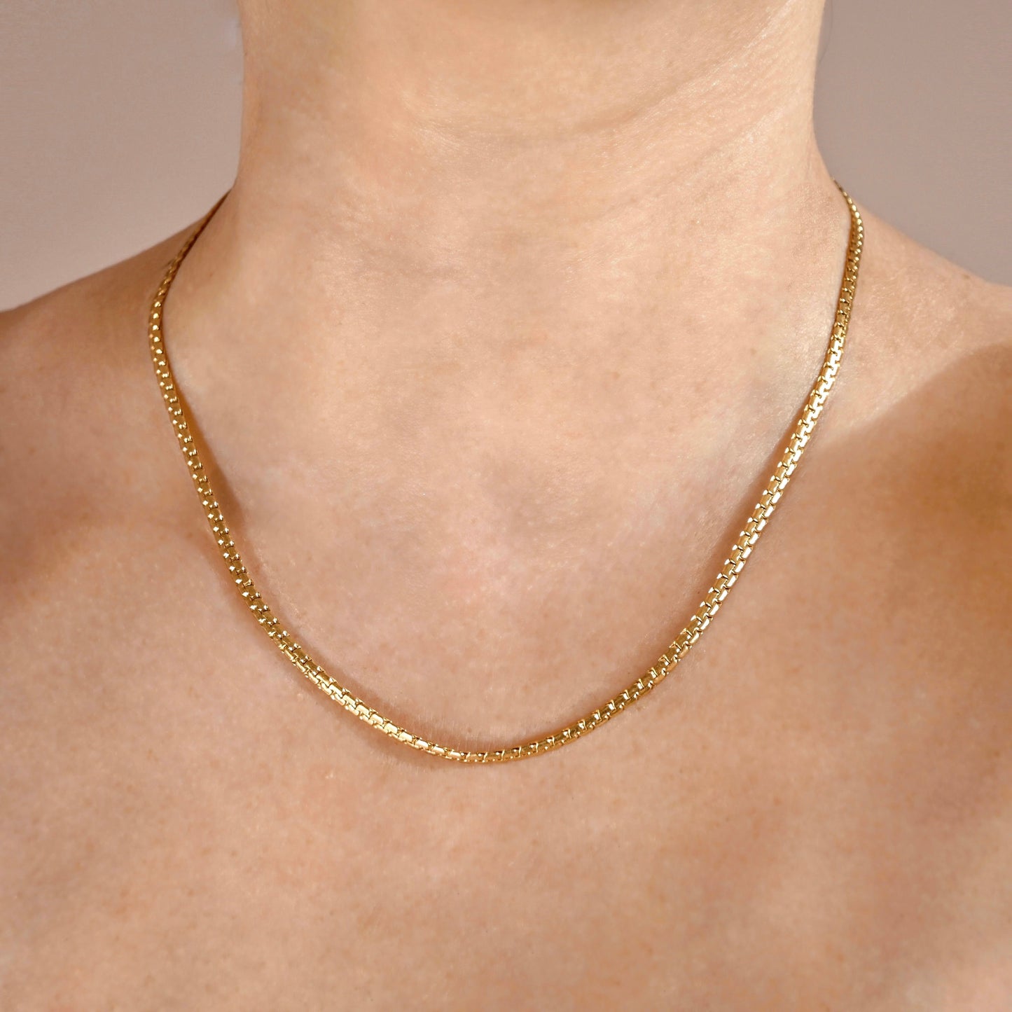 A close-up of a person's neck and upper chest wearing a thick gold snake chain necklace against a soft beige background.