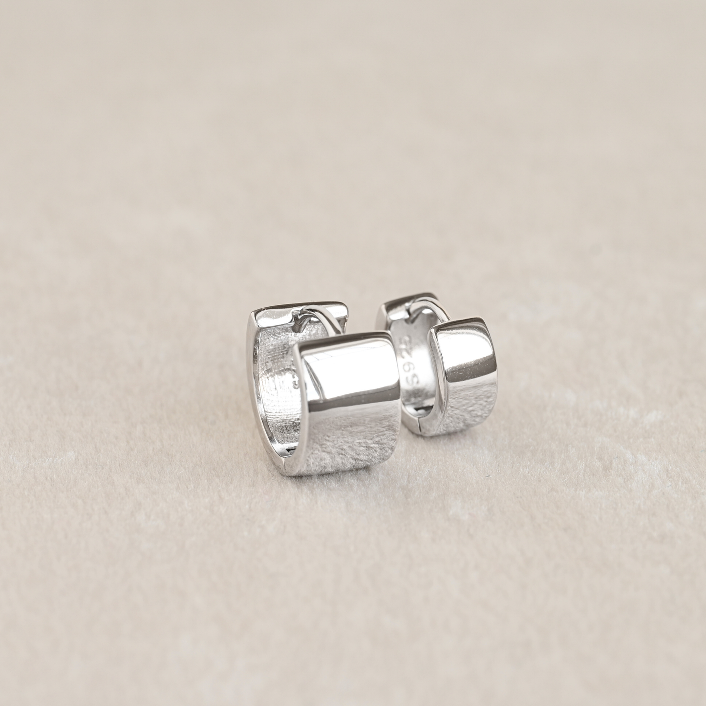 A pair of small silver huggie hoop earrings one medium and wide, one small and thinner, displayed on a neutral beige textured surface.