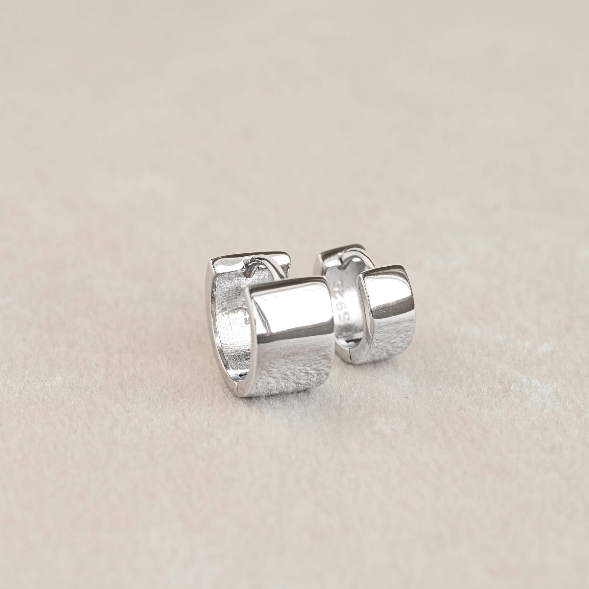 A pair of small silver huggie hoop earrings one medium and wide, one small and thinner, displayed on a neutral beige textured surface.