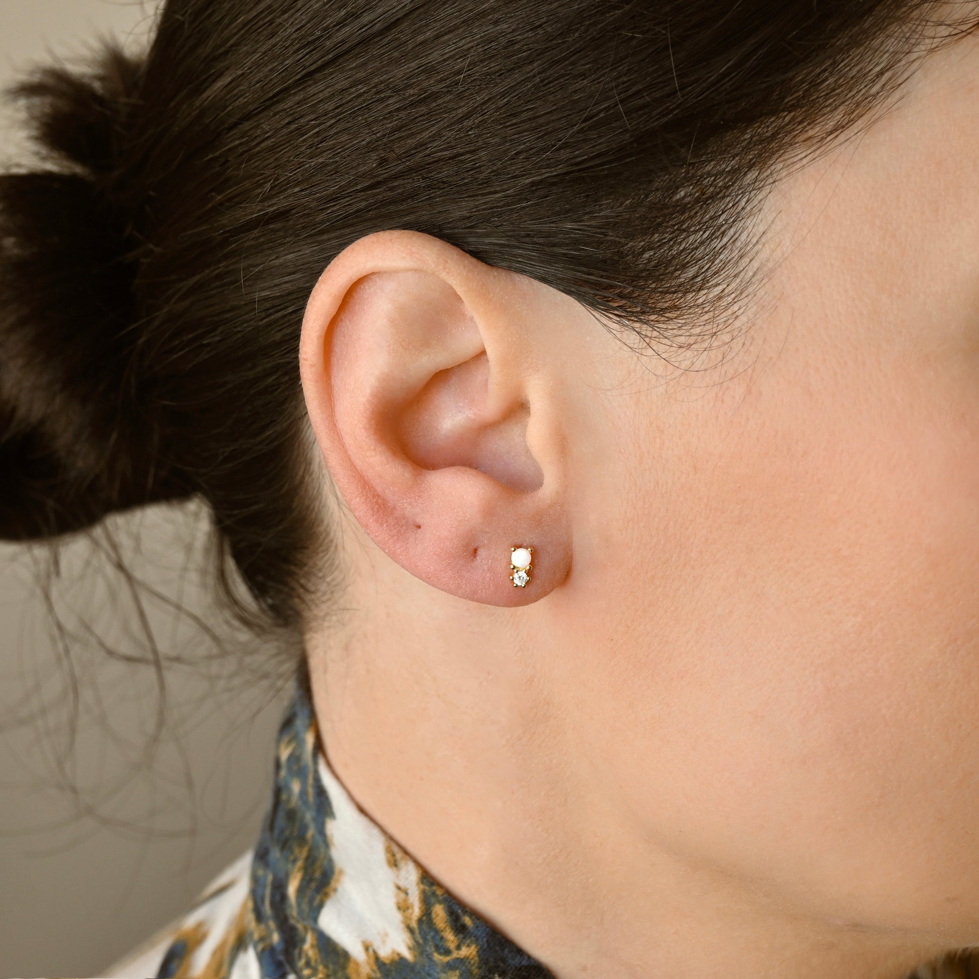 A close-up of a person's ear wearing  small gold stud earring with an opal and cz stones, against a soft neutral background.