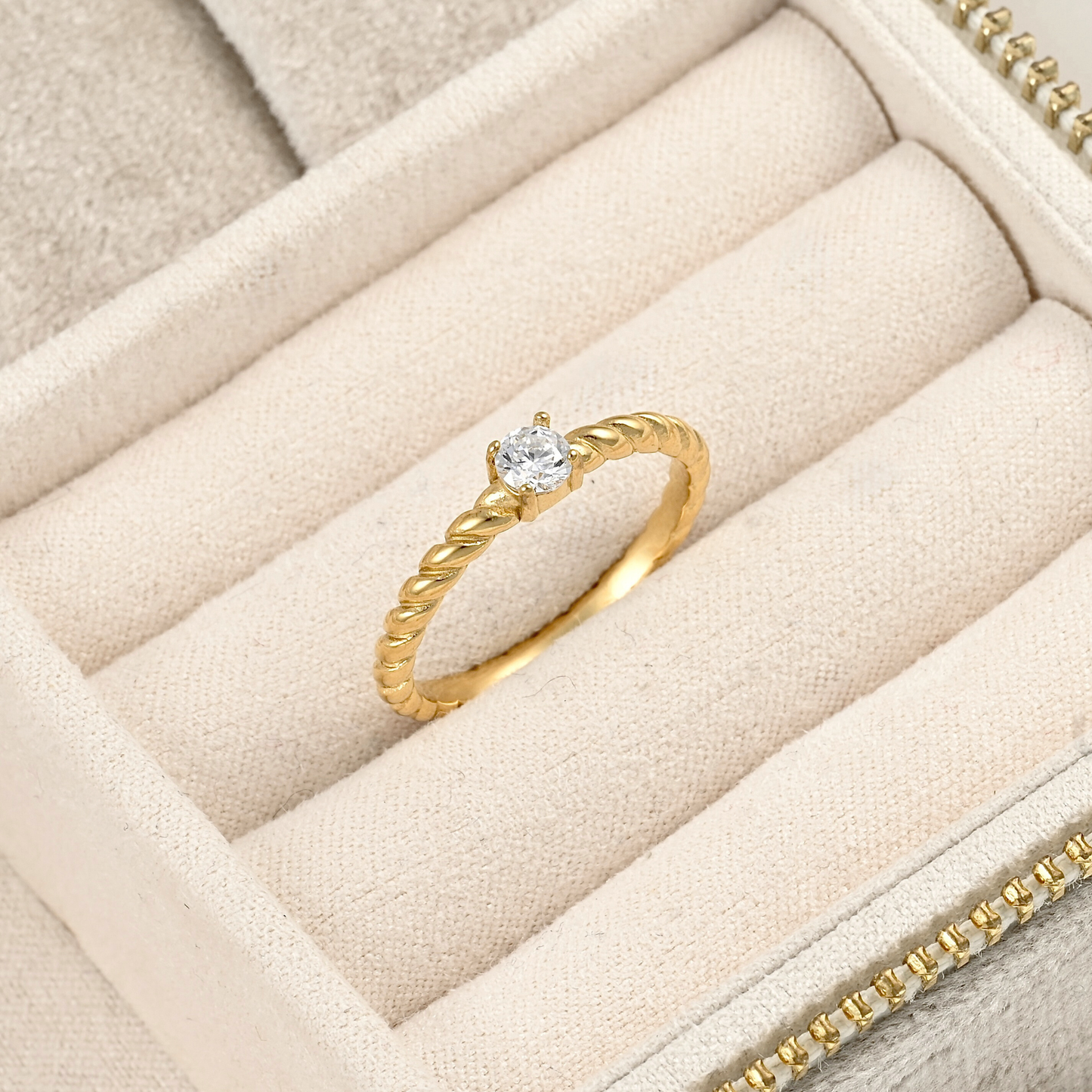 A gold ring with a single round clear gemstone, displayed on a beige velvet jewelry box.
