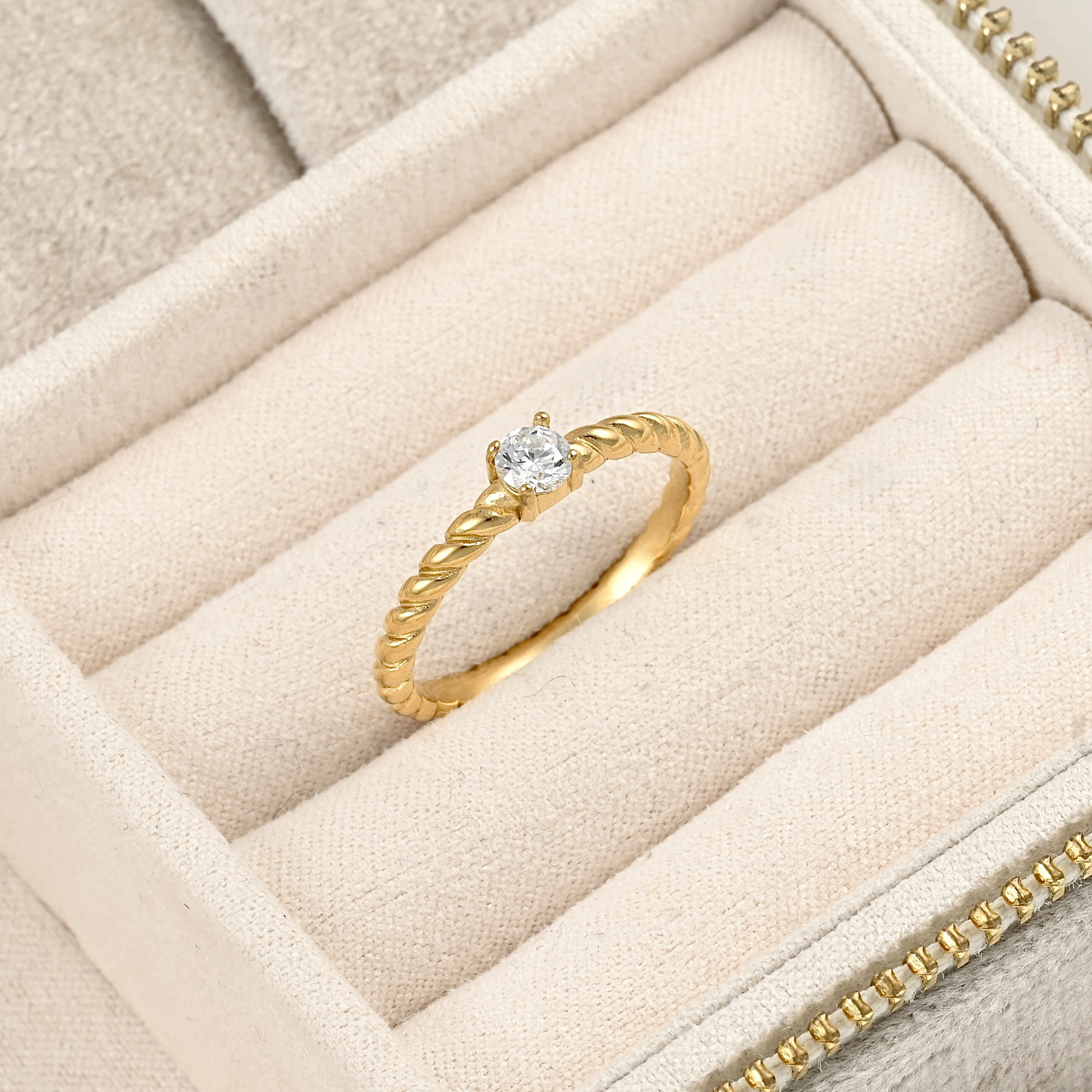 A gold ring with a single round clear gemstone, displayed on a beige velvet jewelry box.