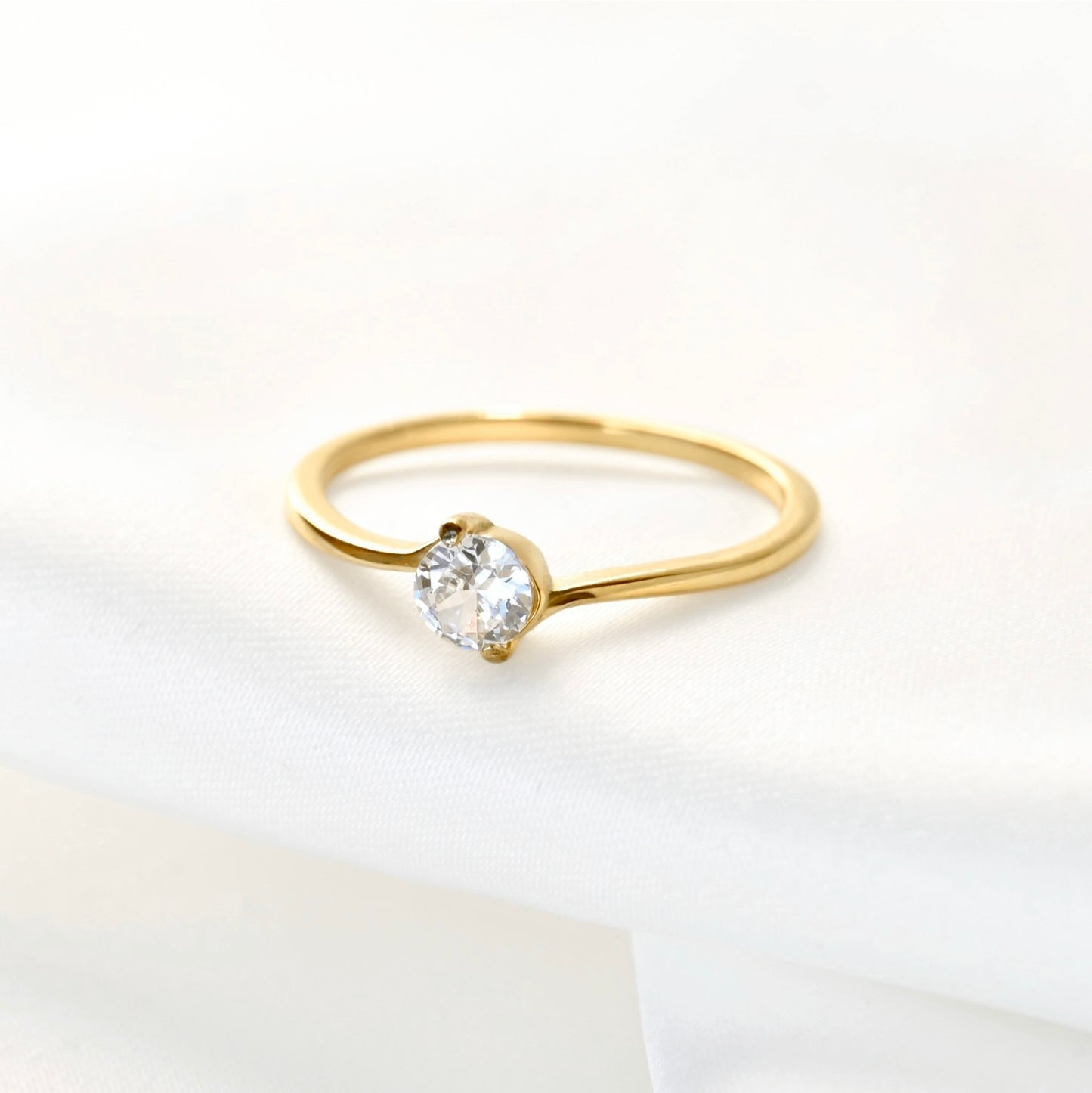 A delicate yellow gold bypass solitaire ring with a round-cut clear gemstone, resting on a soft white fabric surface.