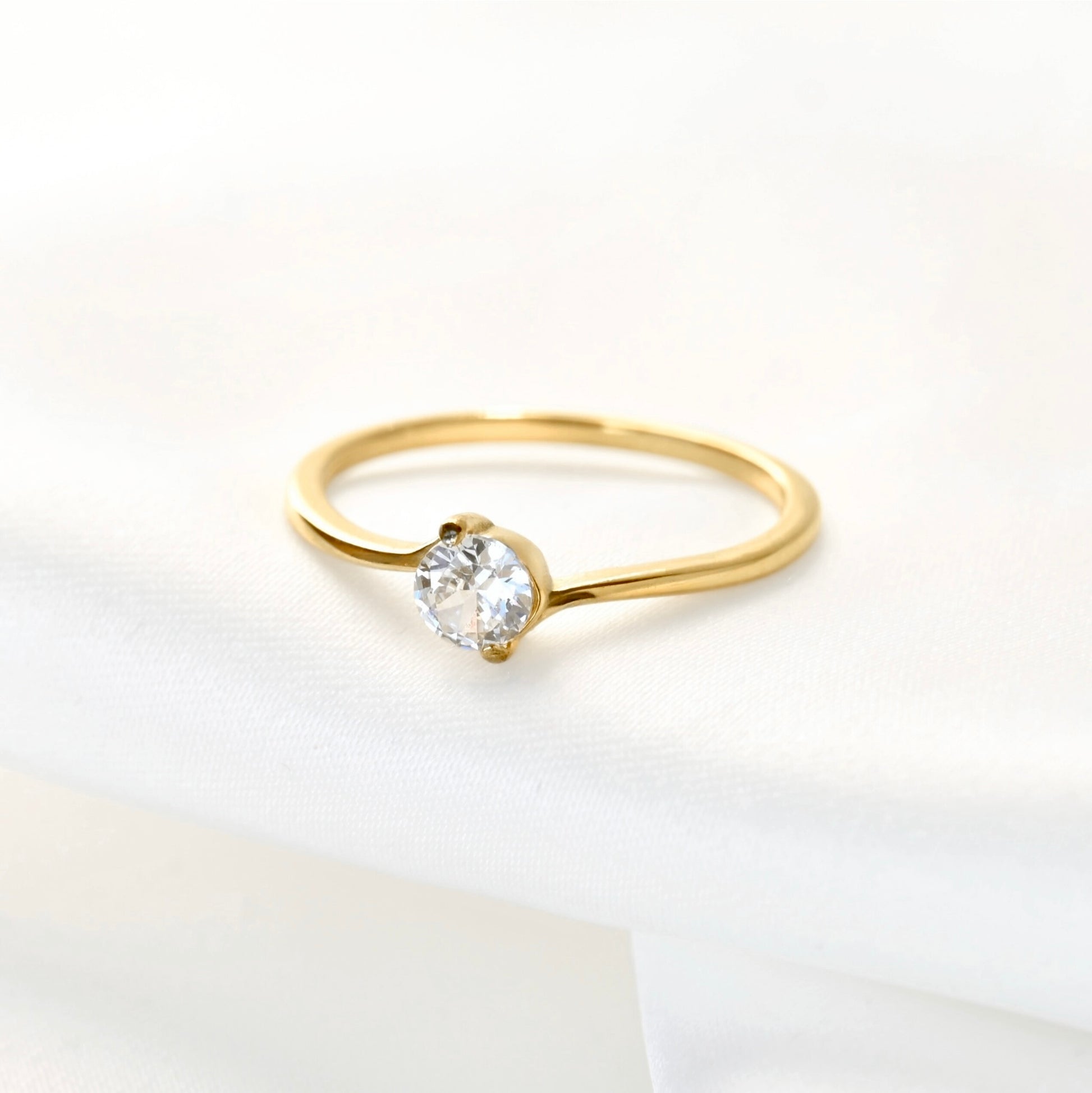 A delicate yellow gold bypass solitaire ring with a round-cut clear gemstone, resting on a soft white fabric surface.