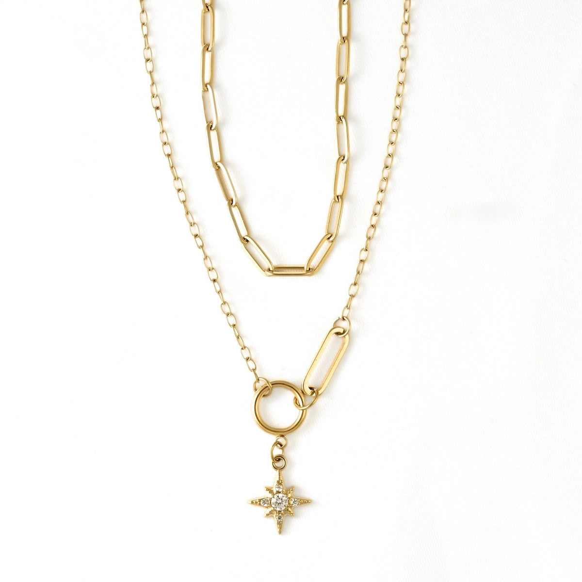 A gold layered necklace featuring a paperclip chain, a link chain, a ring connector, and a small starburst pendant with clear stones, shown on a plain white background.