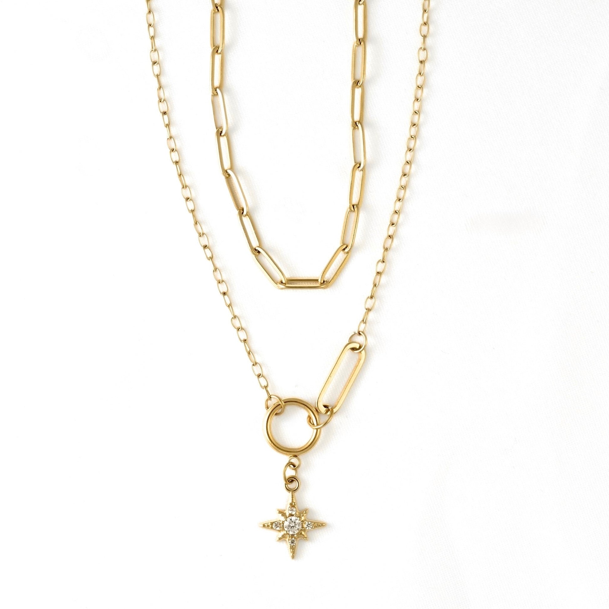 A gold layered necklace featuring a paperclip chain, a link chain, a ring connector, and a small starburst pendant with clear stones, shown on a plain white background.