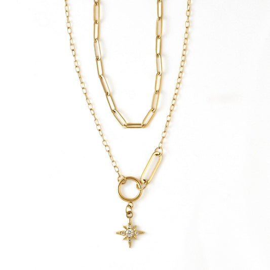 A gold layered necklace featuring a paperclip chain, a link chain, a ring connector, and a small starburst pendant with clear stones, shown on a plain white background.