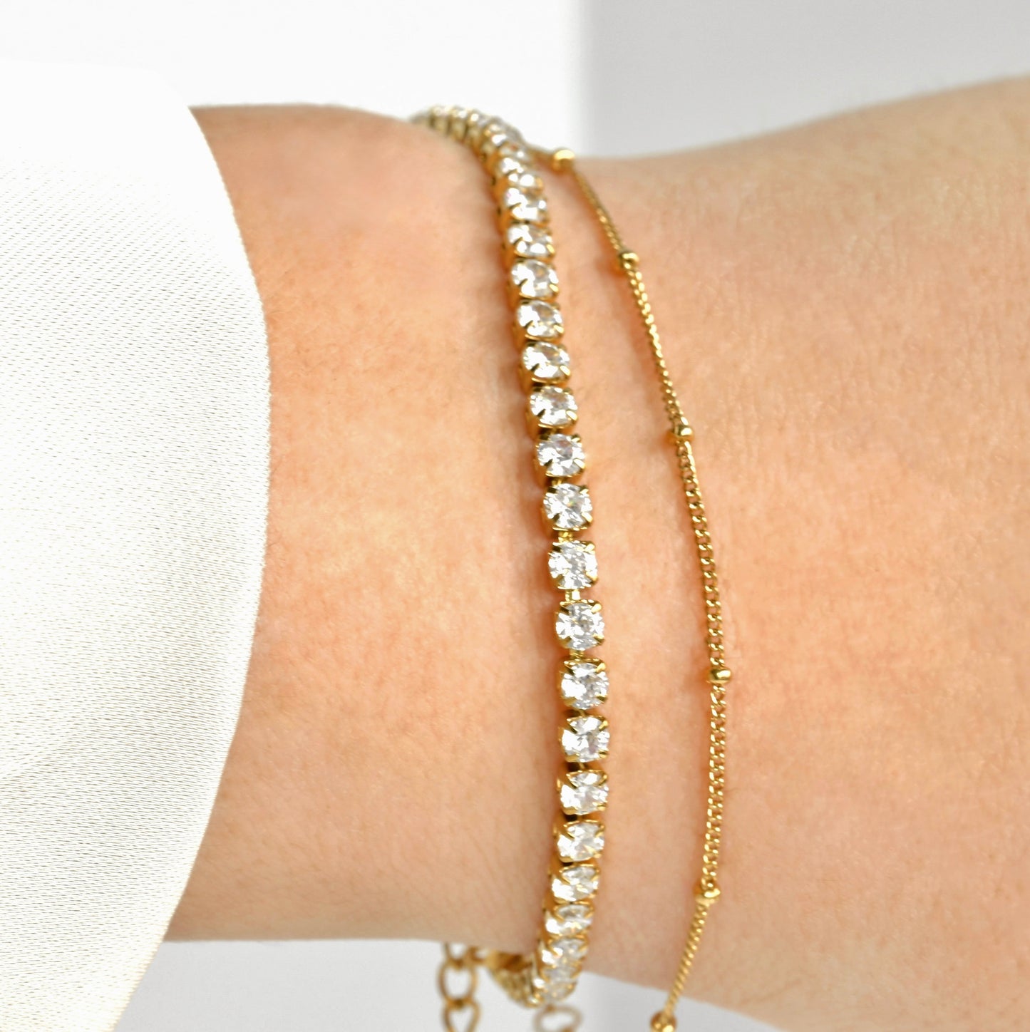 Gold tennis chain bracelet stacked with dainty satellite chain on a wrist against a white background