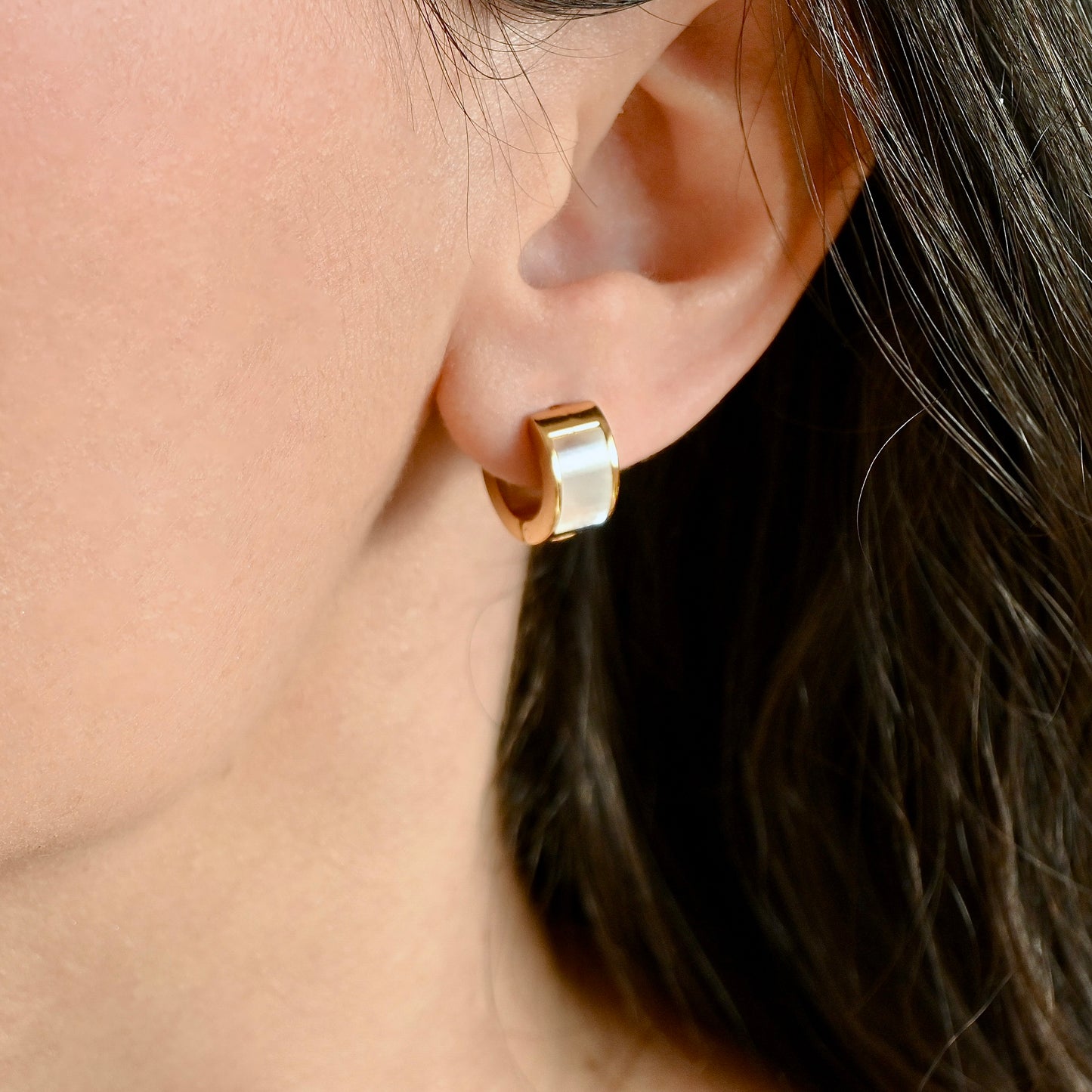 A close-up of a person's ear wearing a small gold huggie hoop earring with a polished, reflective finish.