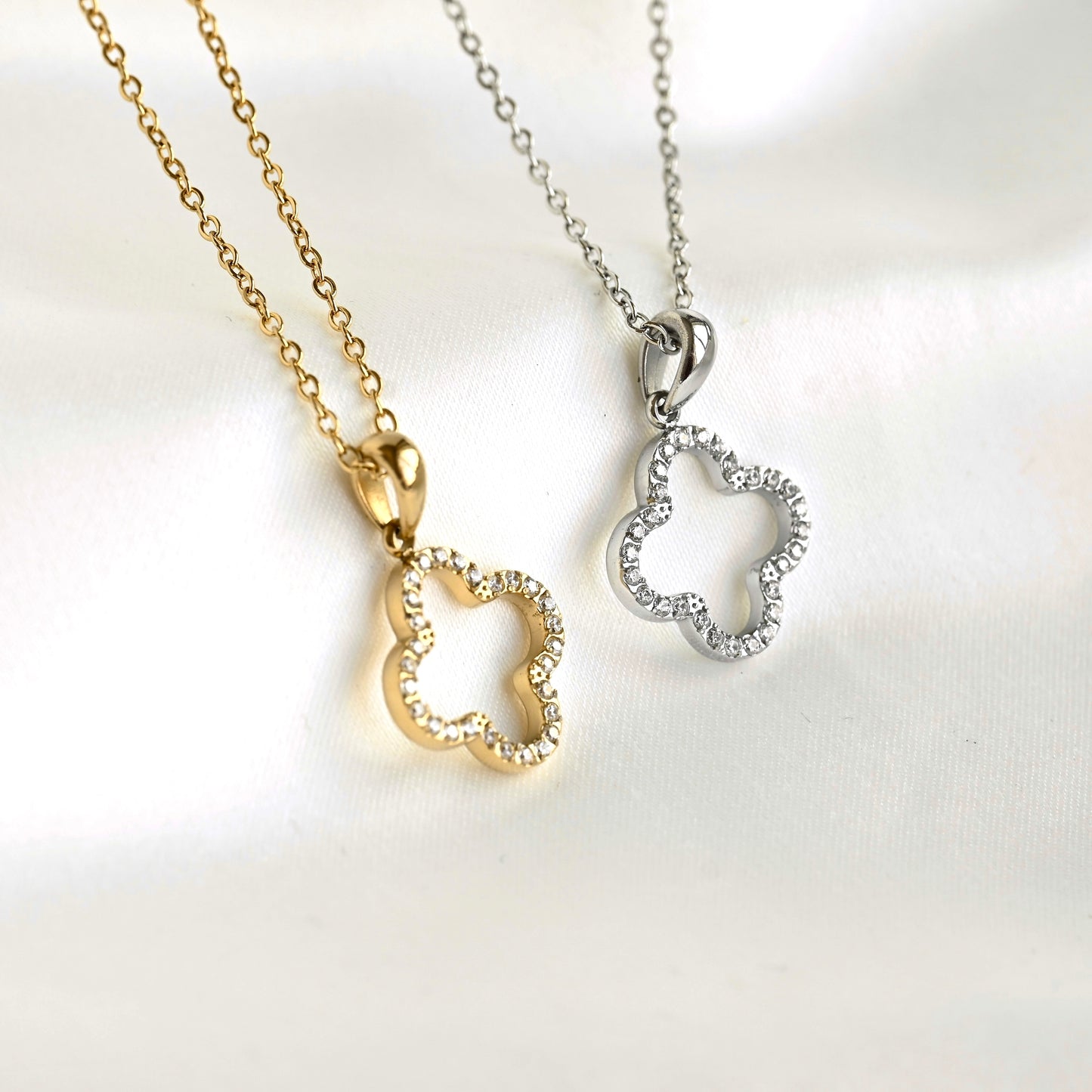 Two clover-shaped pendant necklaces on chains, one gold-tone with clear rhinestones and one silver-tone with clear rhinestones, displayed on a white fabric background.
