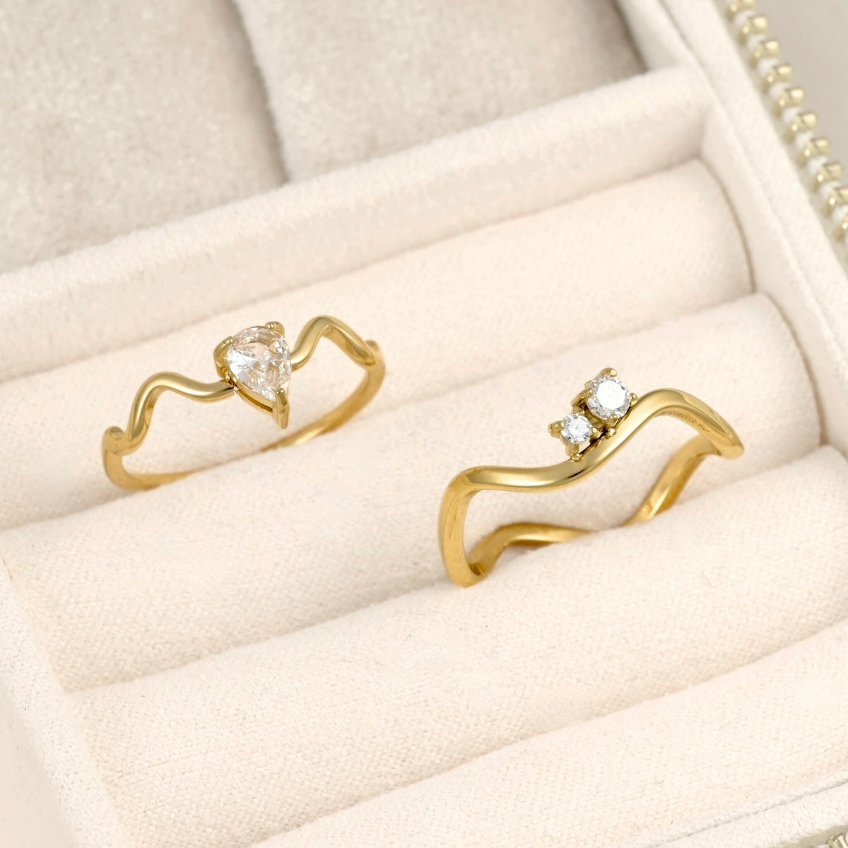 Two gold rings displayed in a cream-colored jewelry box. One ring features a wavy band with a small clear heart-shaped gemstone, and the other features a wavy band with two small clear round-cut gemstones.