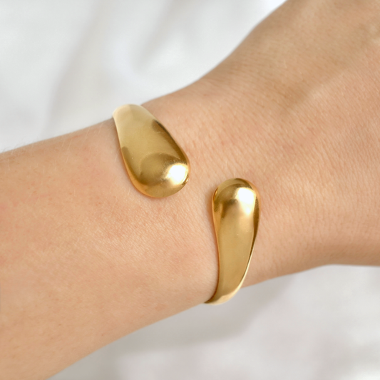 A close-up of a person's wrist wearing a minimalist, open cuff bracelet in polished yellow gold, set against a soft, out-of-focus white background.