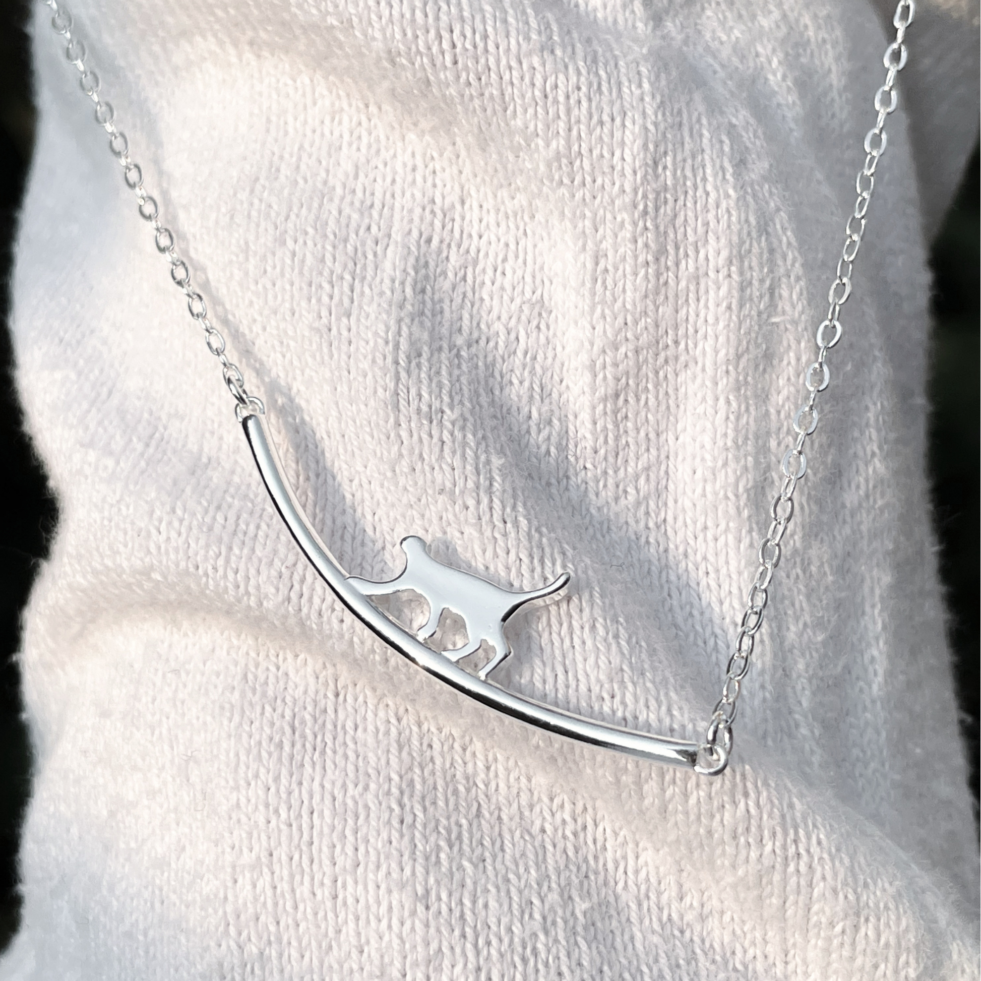 Silver cat necklace with a curved bar design on a white sweater under direct sunlight