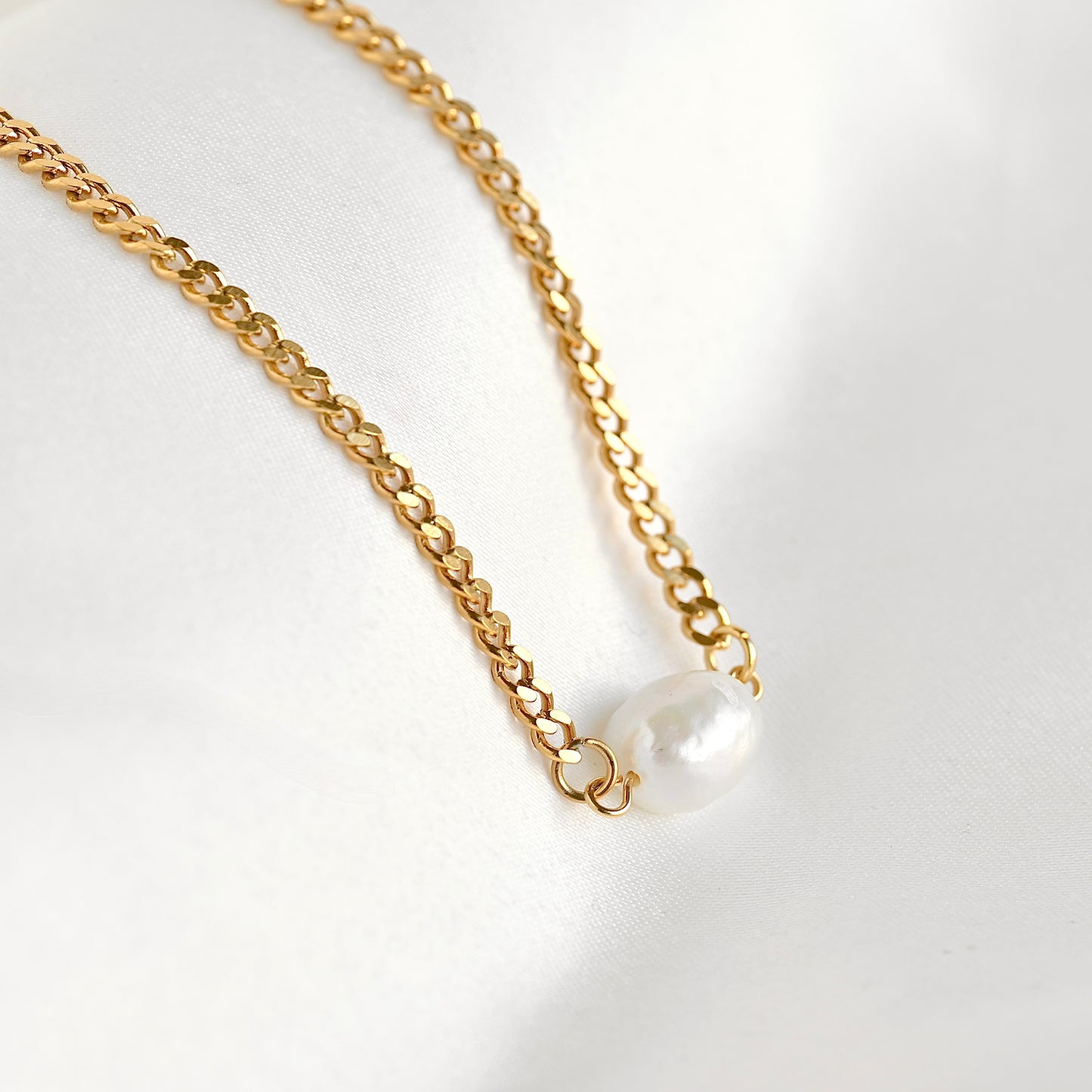 A gold curb chain necklace featuring a single baroque pearl pendant, displayed on a soft white fabric background.