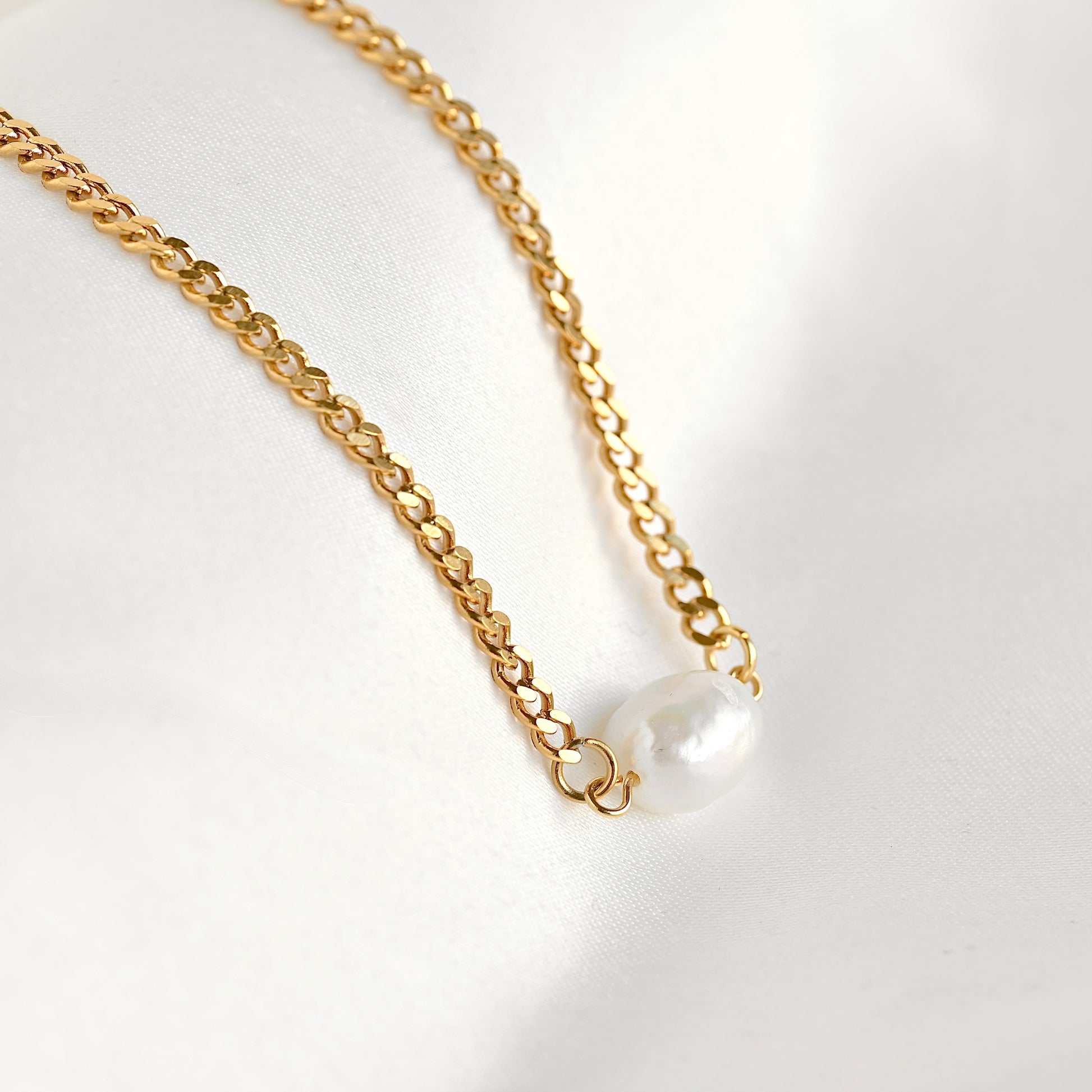 A gold curb chain necklace featuring a single baroque pearl pendant, displayed on a soft white fabric background.