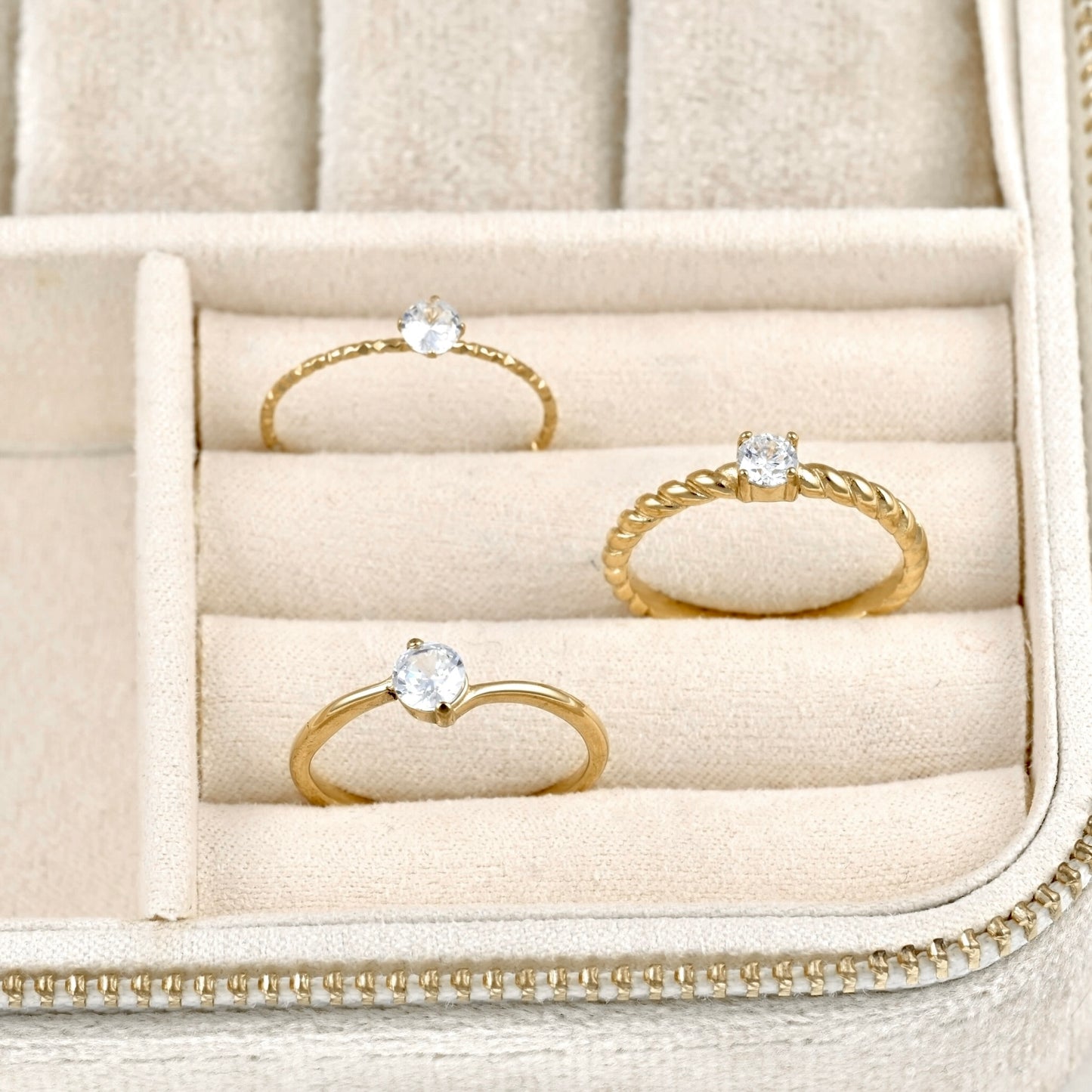 Three gold rings with clear gemstones displayed in a beige jewelry box.
