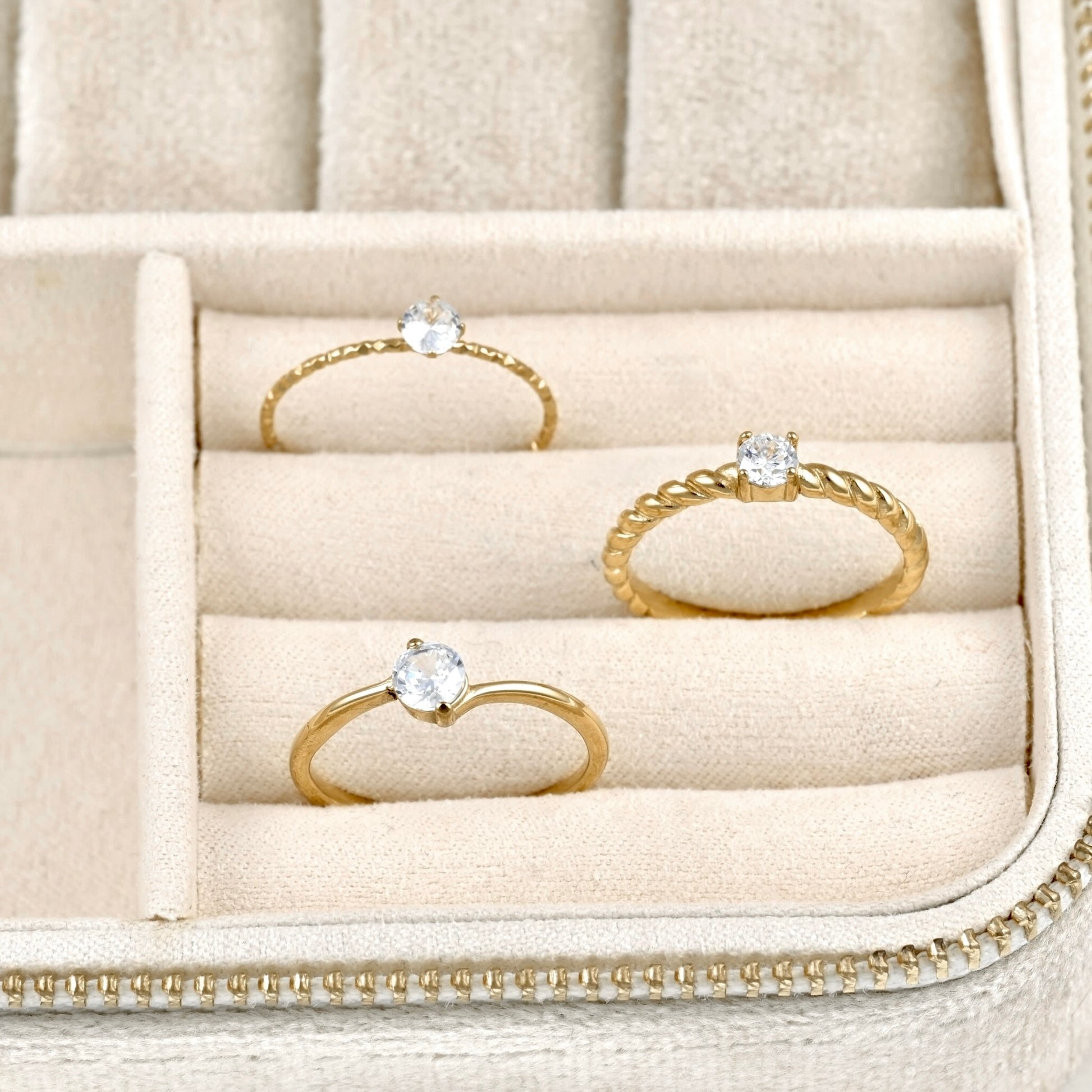 Three gold rings with clear gemstones displayed in a beige jewelry box.