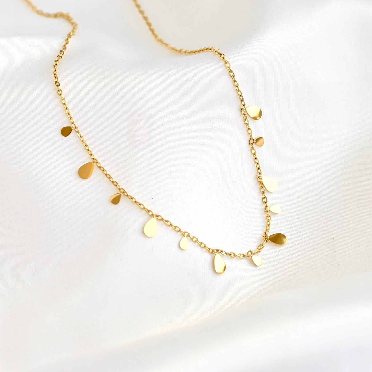 A delicate gold chain necklace with small, irregular flat gold leaf-like pendants, displayed on a soft white fabric background.