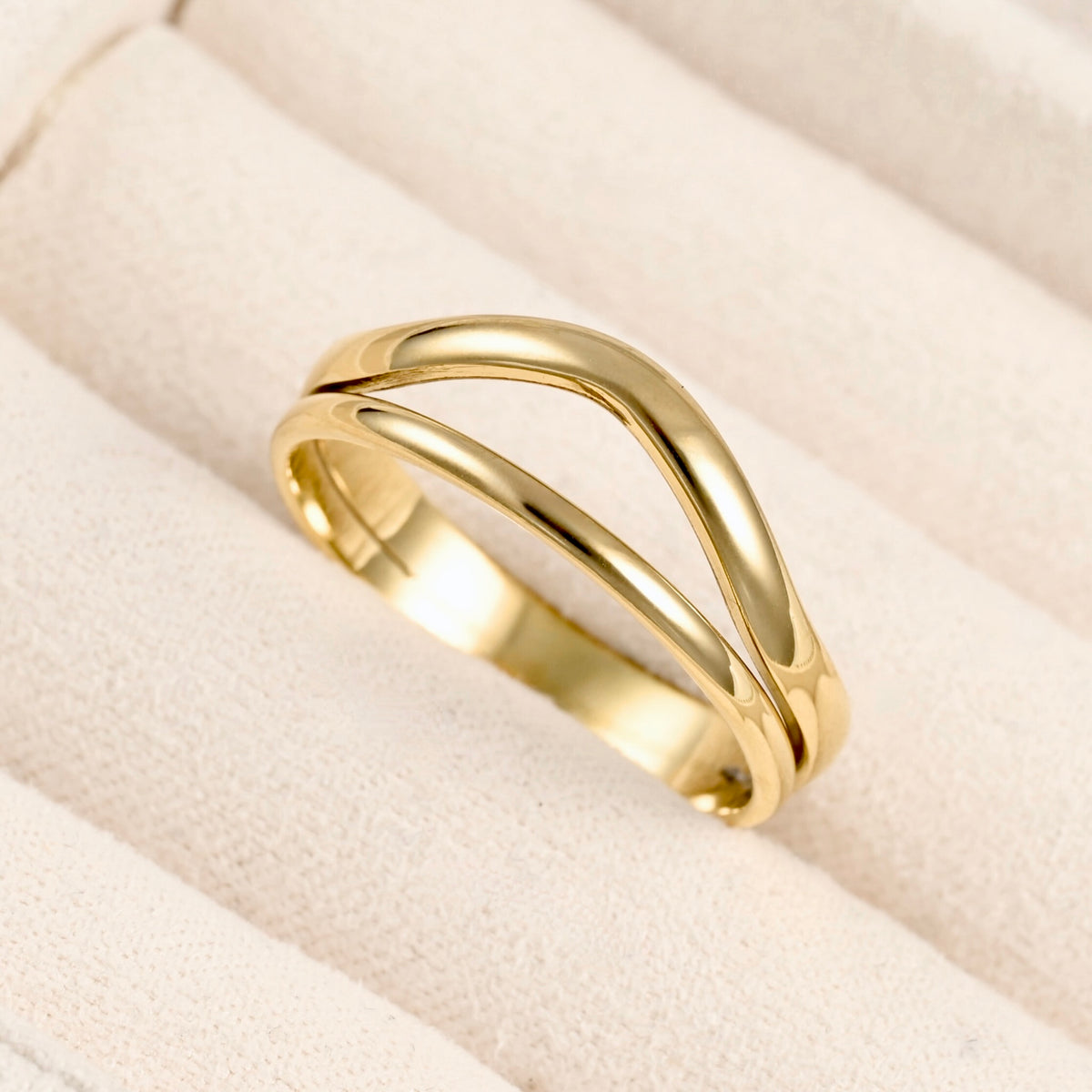 A polished yellow gold ring with a double, flowing wave design, displayed on a textured cream-colored ring box.