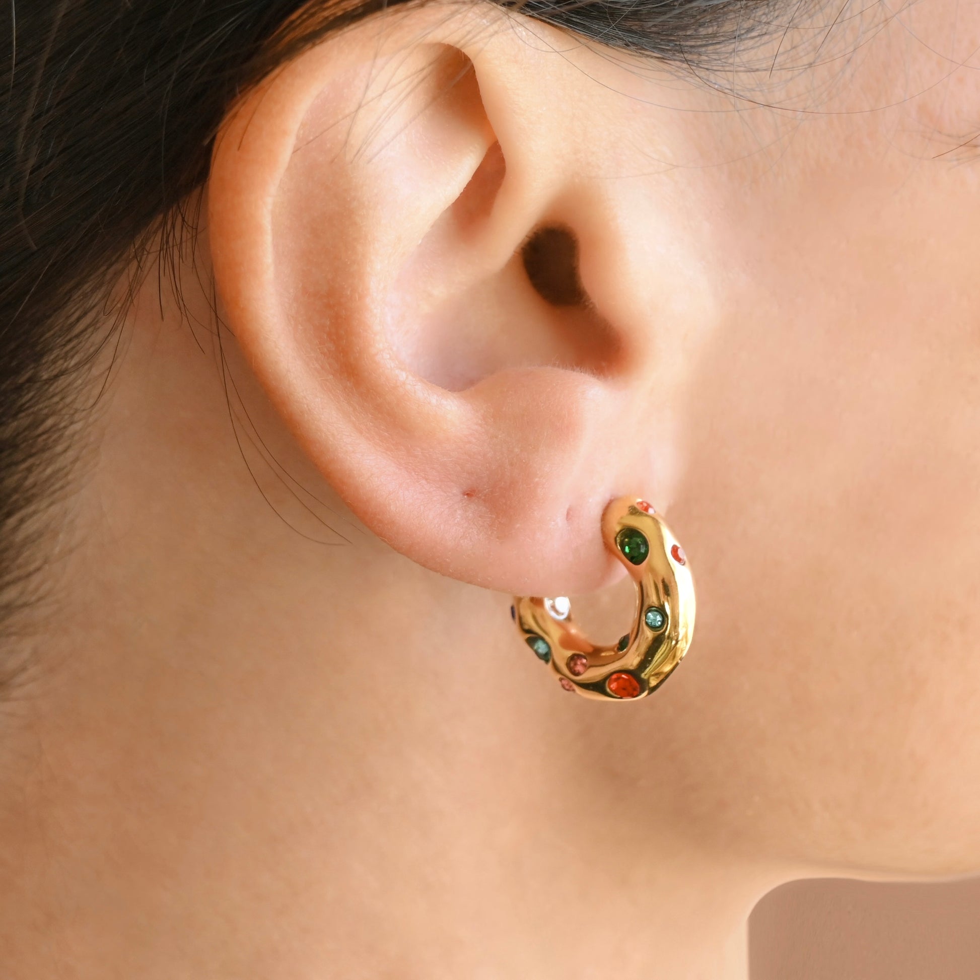 A close-up of a person's ear wearing a medium chunky gold hoop earring decorated with colorful small clear stones of different sizes.