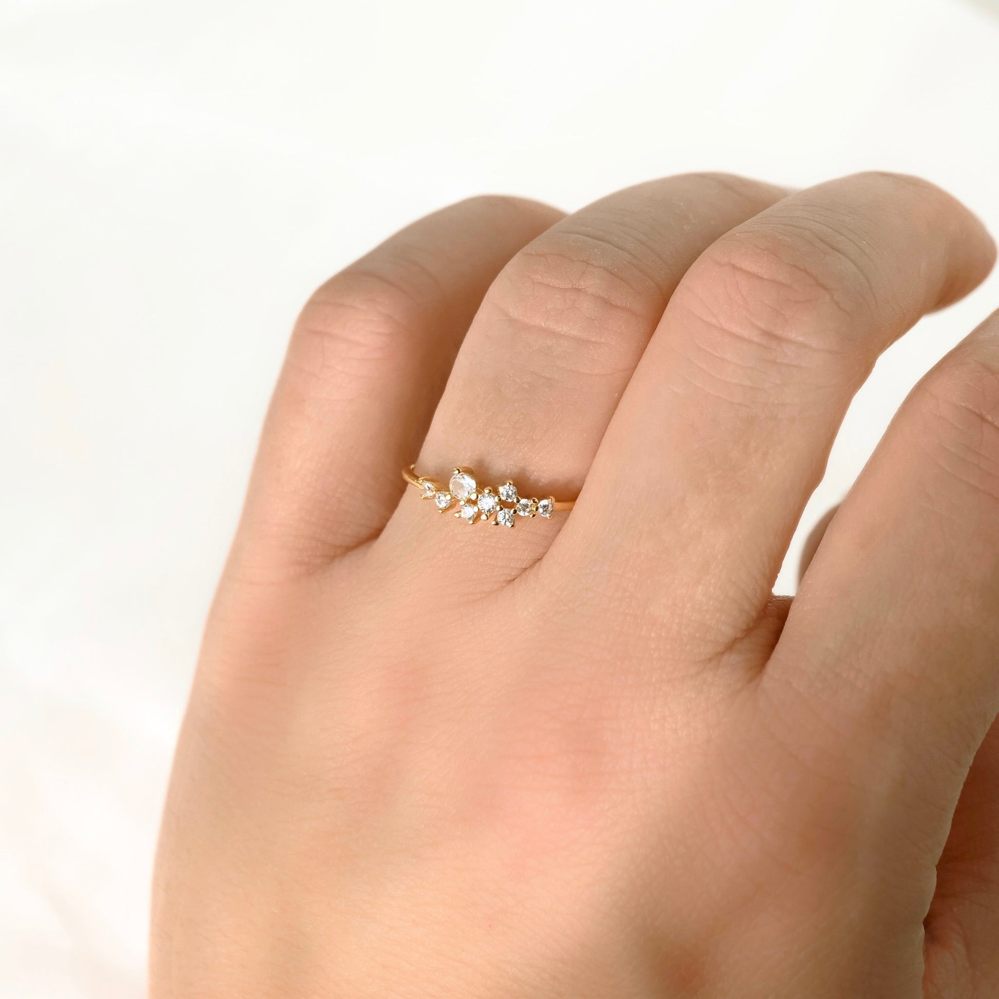 A delicate gold ring with a small cluster of clear stones on the middle finger of a hand, photographed against a soft white background.