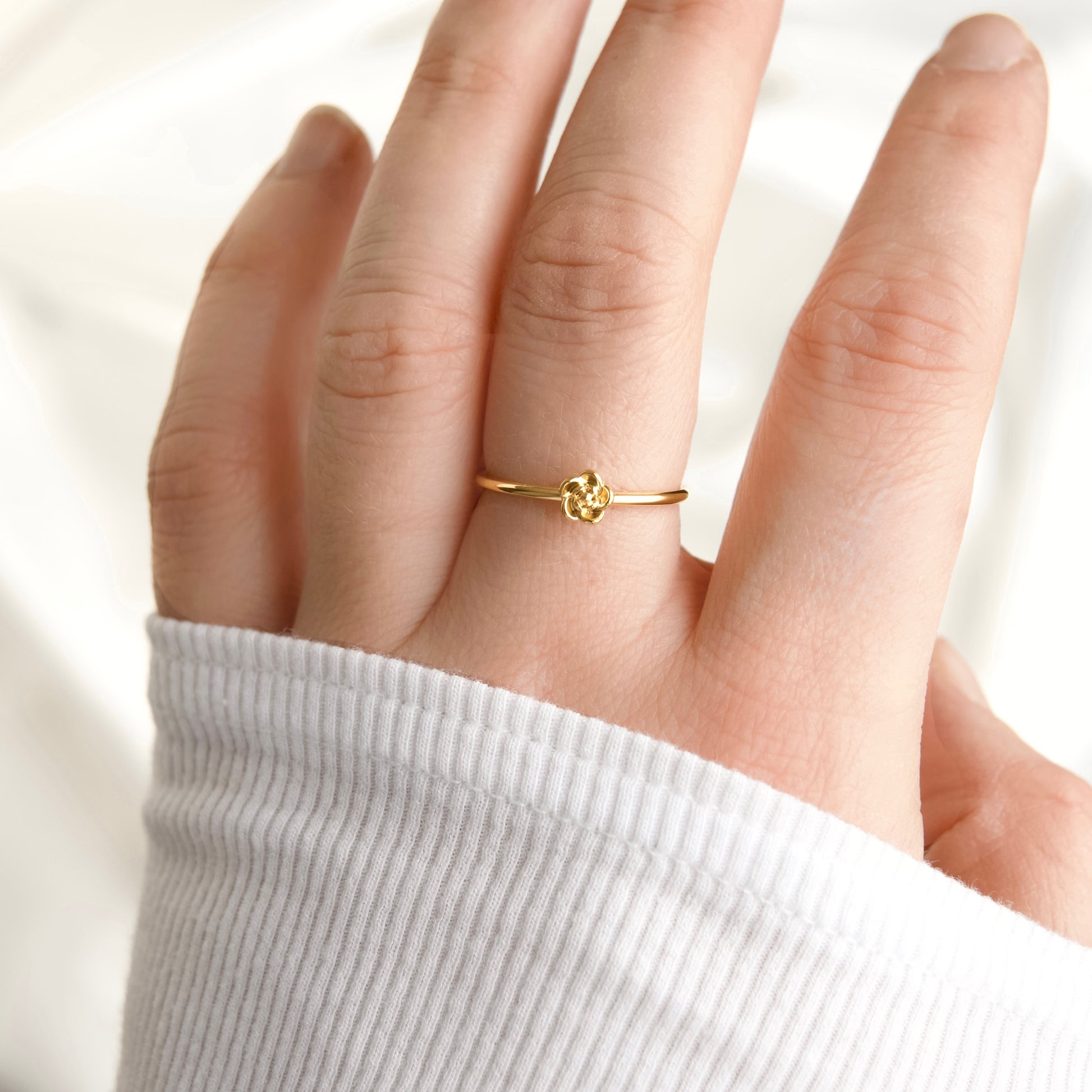 A delicate gold ring with a small flower-like center, worn on a person's ring finger. The hand is wearing a white ribbed long-sleeve top, and the background is a soft, out-of-focus white fabric.